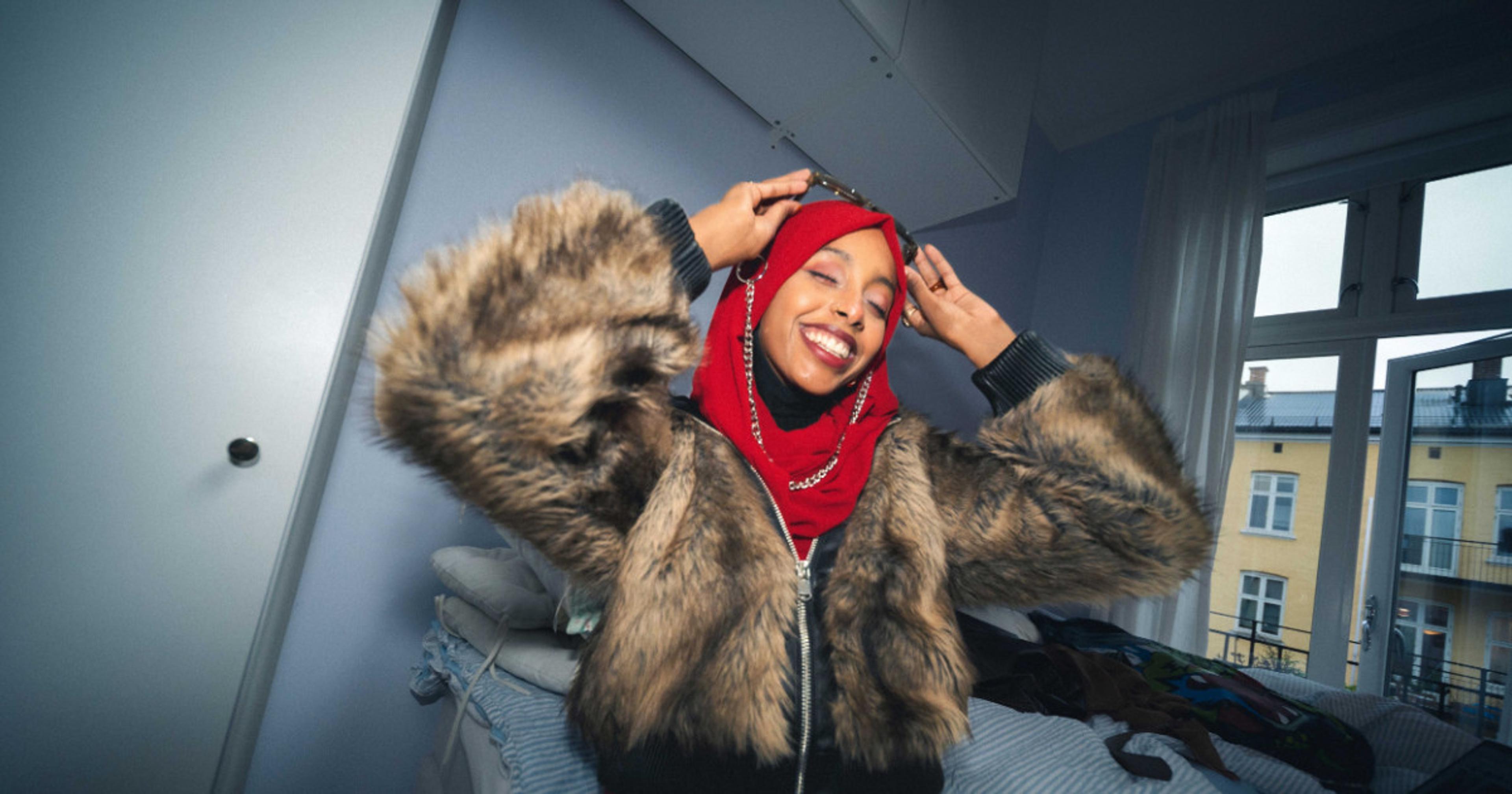 Smilende ung kvinne med rød hijab og tykk fuskepelsjakke står i et lyst soverom og justerer solbriller på hodet.