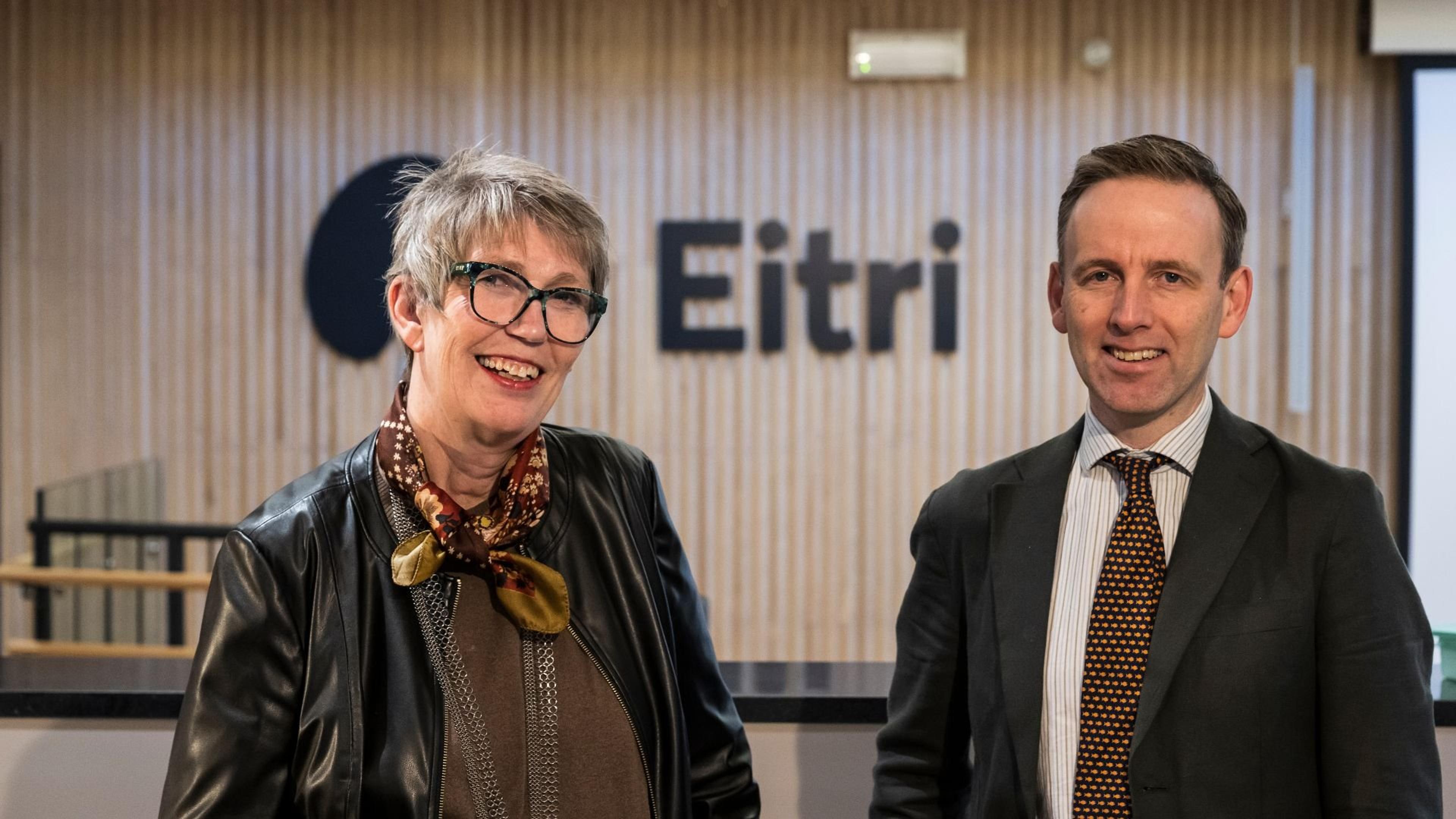 En smilende kvinne med briller og en mann i dress står foran en trevegg med Eitri-logoen.