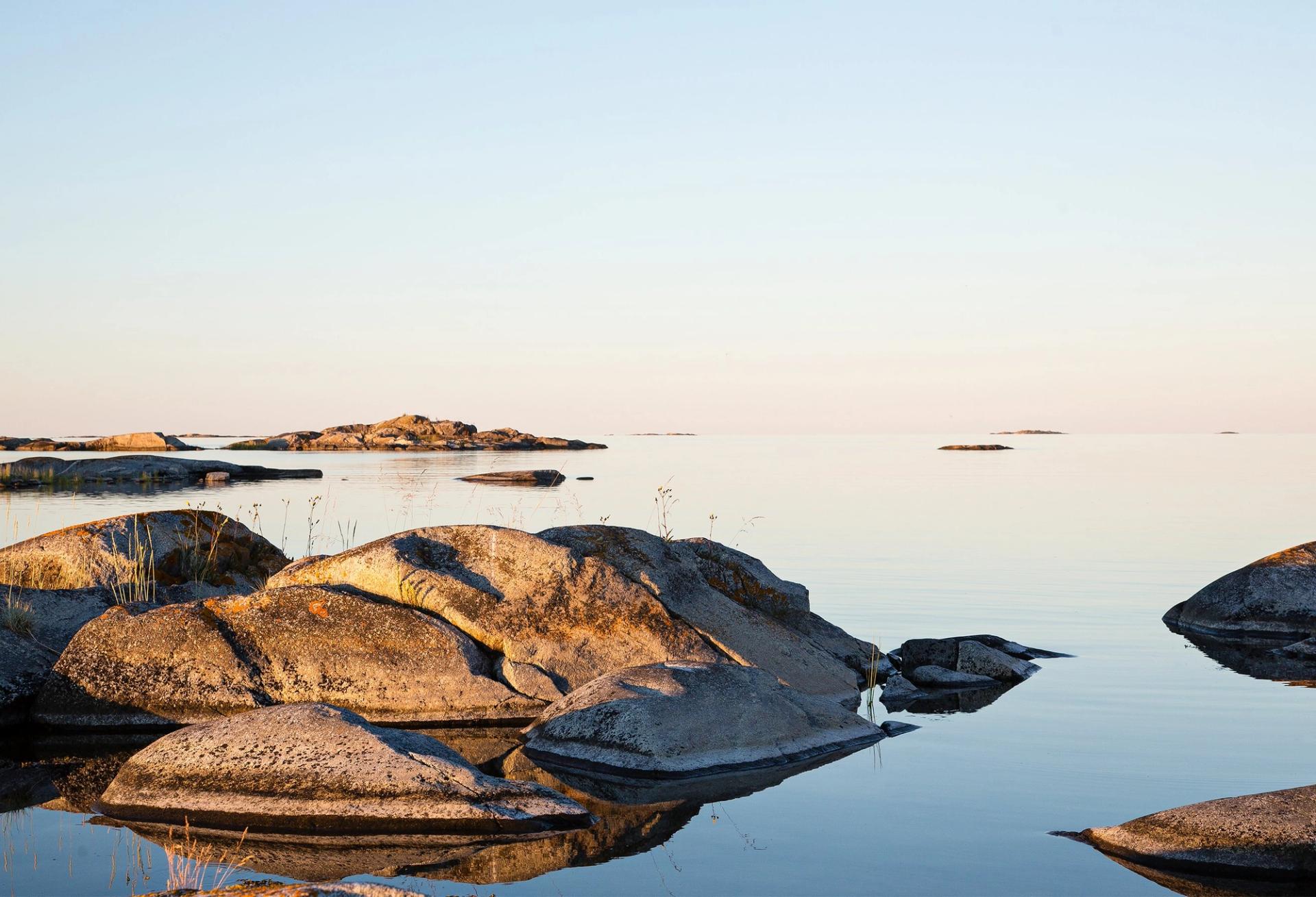 Rolig kystlandskap med glatte svaberg som speiler seg i stille vann under en lys, klar himmel ved solnedgang eller soloppgang.