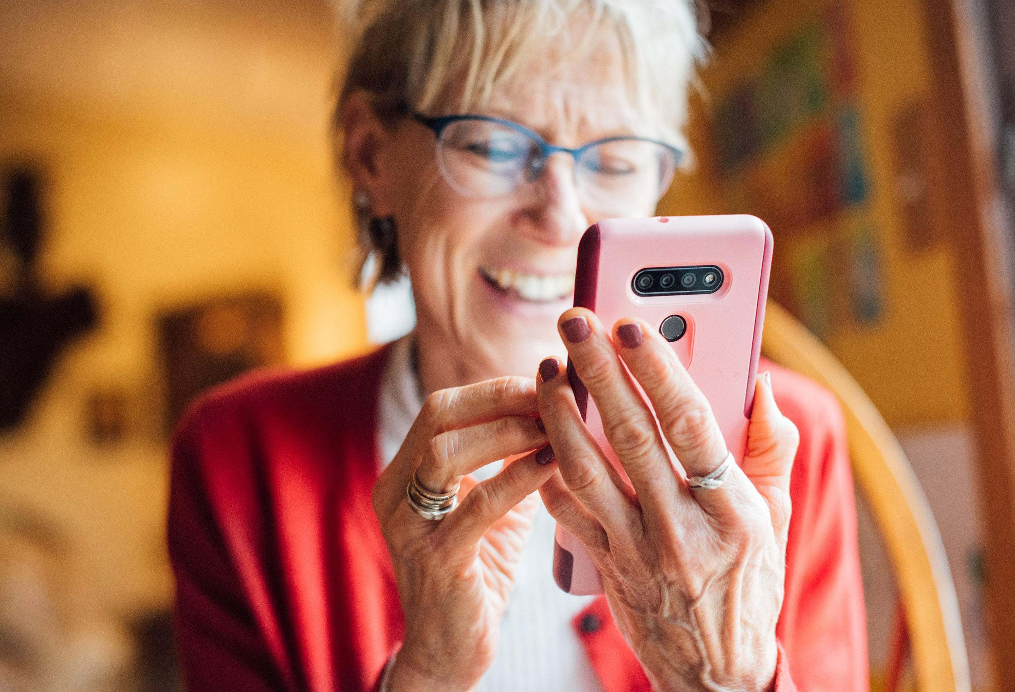 Eldre kvinne med briller og rød jakke smiler mens hun holder og bruker en smarttelefon med rosa deksel innendørs.