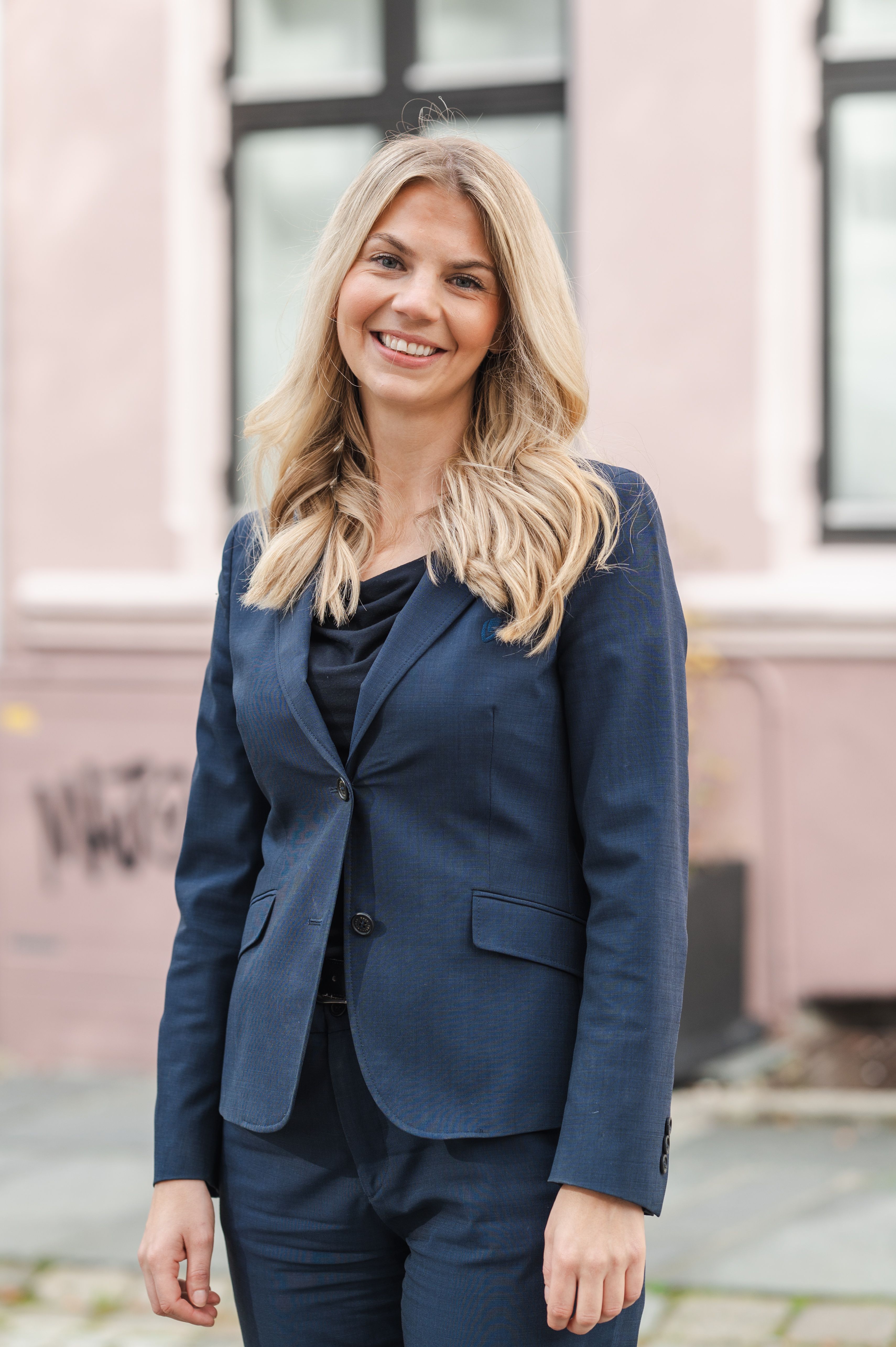 Rådgiver Kristine Helene Steiner. Smilende kvinne med langt, blondt hår i mørkeblå dress står foran en rosa bygning.