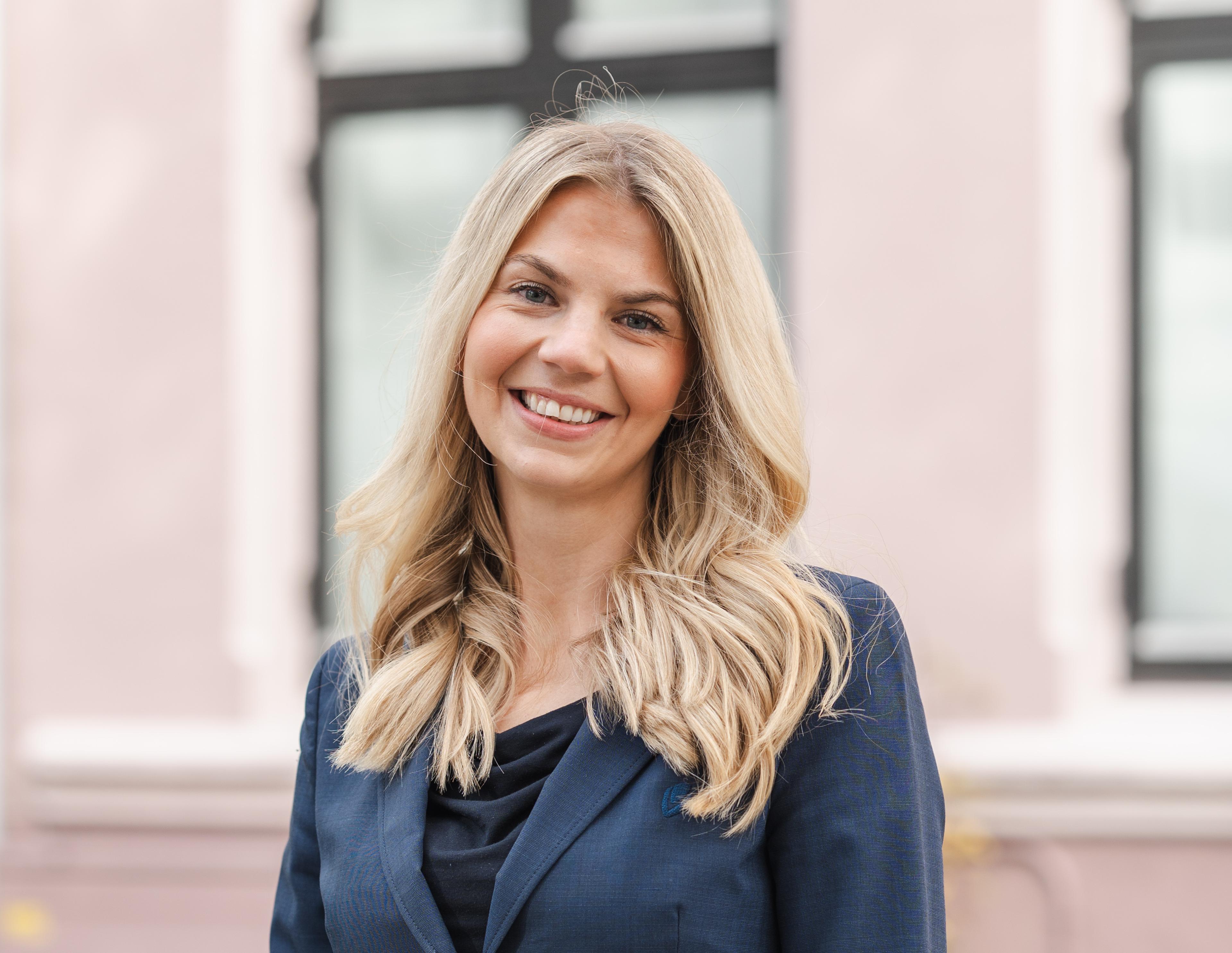 Smilende kvinne med langt blondt hår i mørk blå blazer står utendørs i en bygårdsgate med uskarpe, fargerike hus og grønne planter i bakgrunnen.