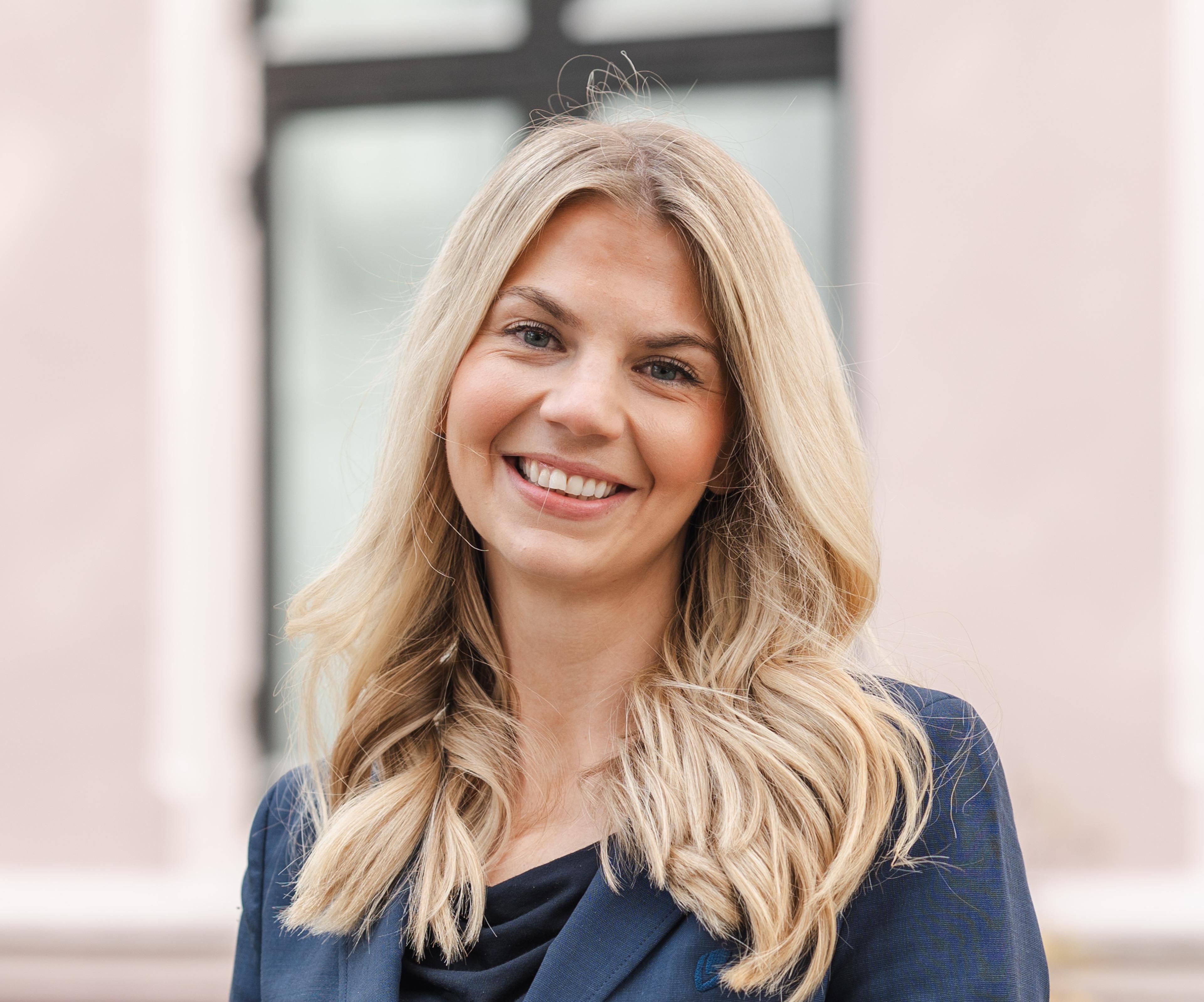 Rådgiver Kristine Helene Steiner. Smilende kvinne med langt, blondt hår i mørkeblå dress står foran en rosa bygning.