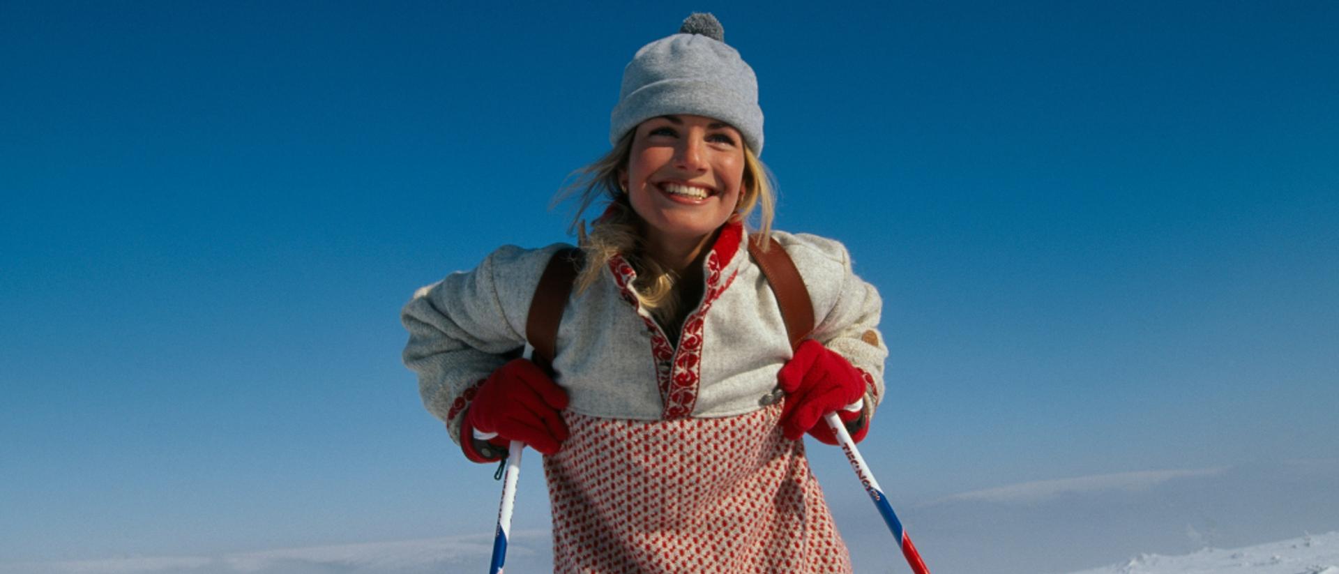 Smilende person på ski i snødekt landskap, kledd i tradisjonell ullgenser, rødvotter og grå lue under klar blå himmel.