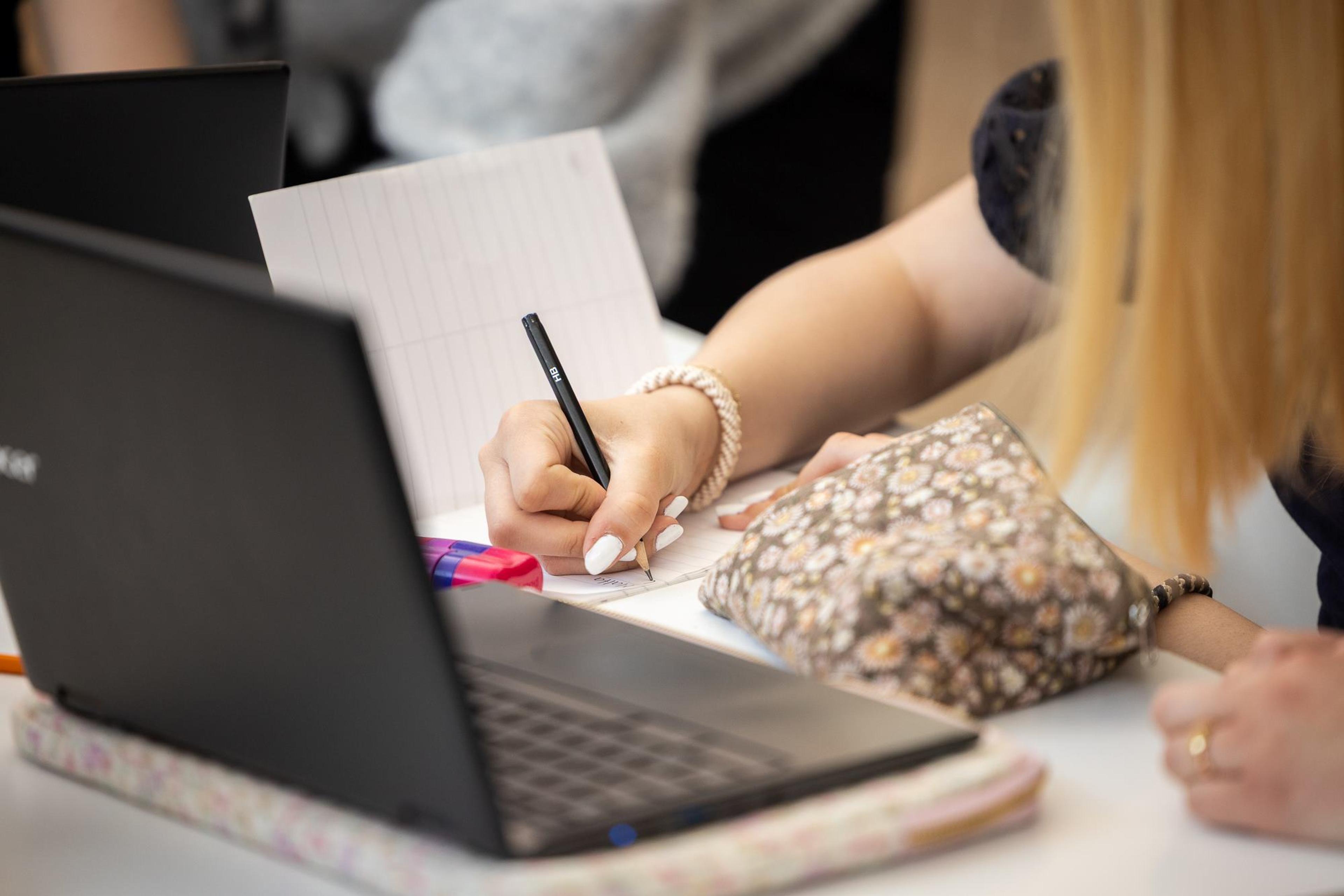 Person sitter ved PC og penn i hånden og skriver på papir