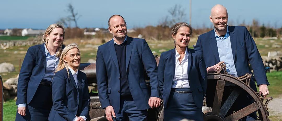 Fem smilende personer i mørke blå dresser står utendørs foran et gammelt hjul, med åpent landskap og gårdsbygninger i bakgrunnen.