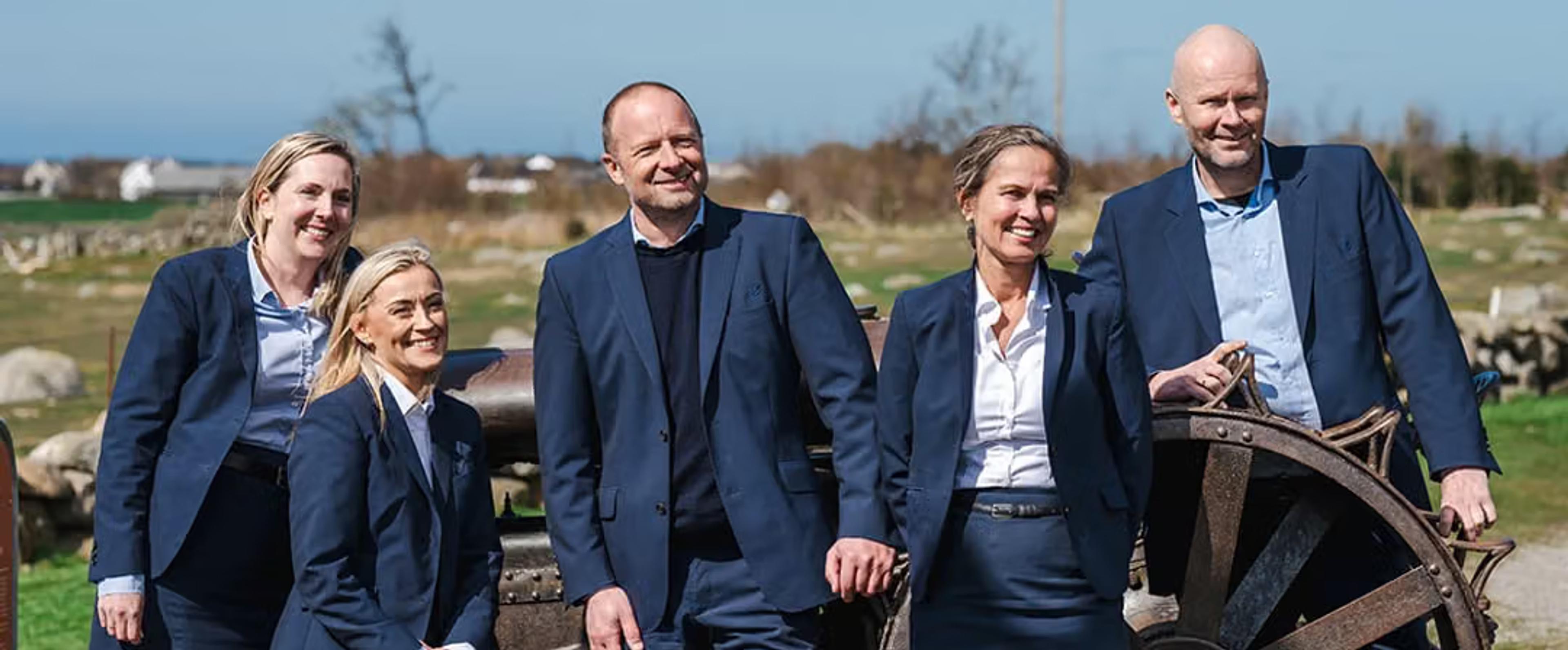 Fem smilende personer i mørke blå dresser står utendørs foran et gammelt hjul, med åpent landskap og gårdsbygninger i bakgrunnen.