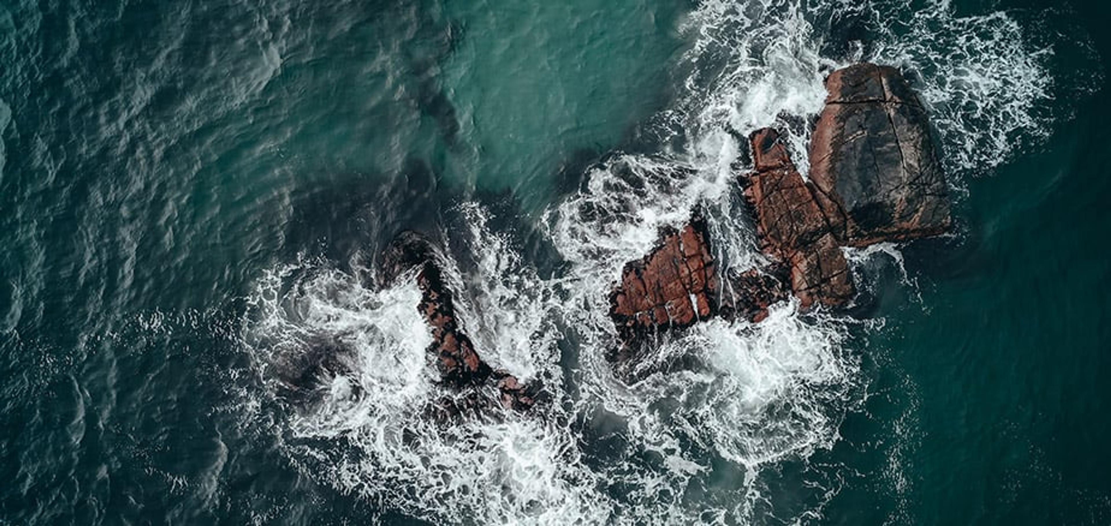 Luftfoto av mørkeblått hav som slår inn mot rødlige klipper, med hvitt skum som bruser rundt steinene.
