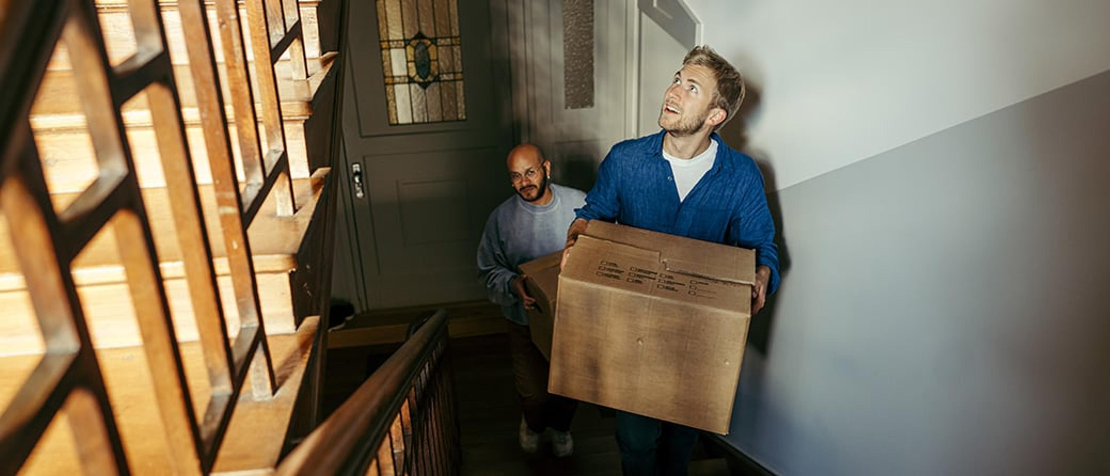 To menn bærer en stor pappeske opp en trapp i en oppgang, mens den fremste ser oppover mot etasjen over.