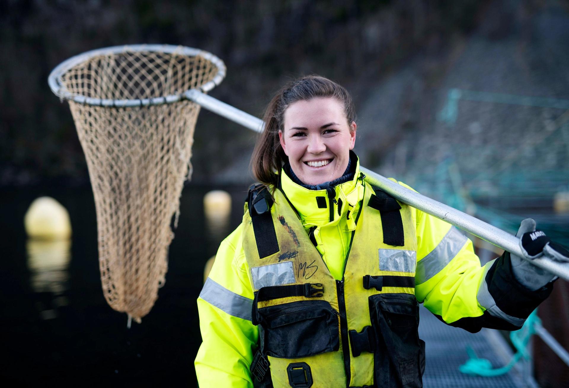 En kvinne i en neongul arbeidsjakke sitter bak en glassrute og ser konsentrert ut til siden. Refleksjoner i glasset viser omgivelsene rundt henne.