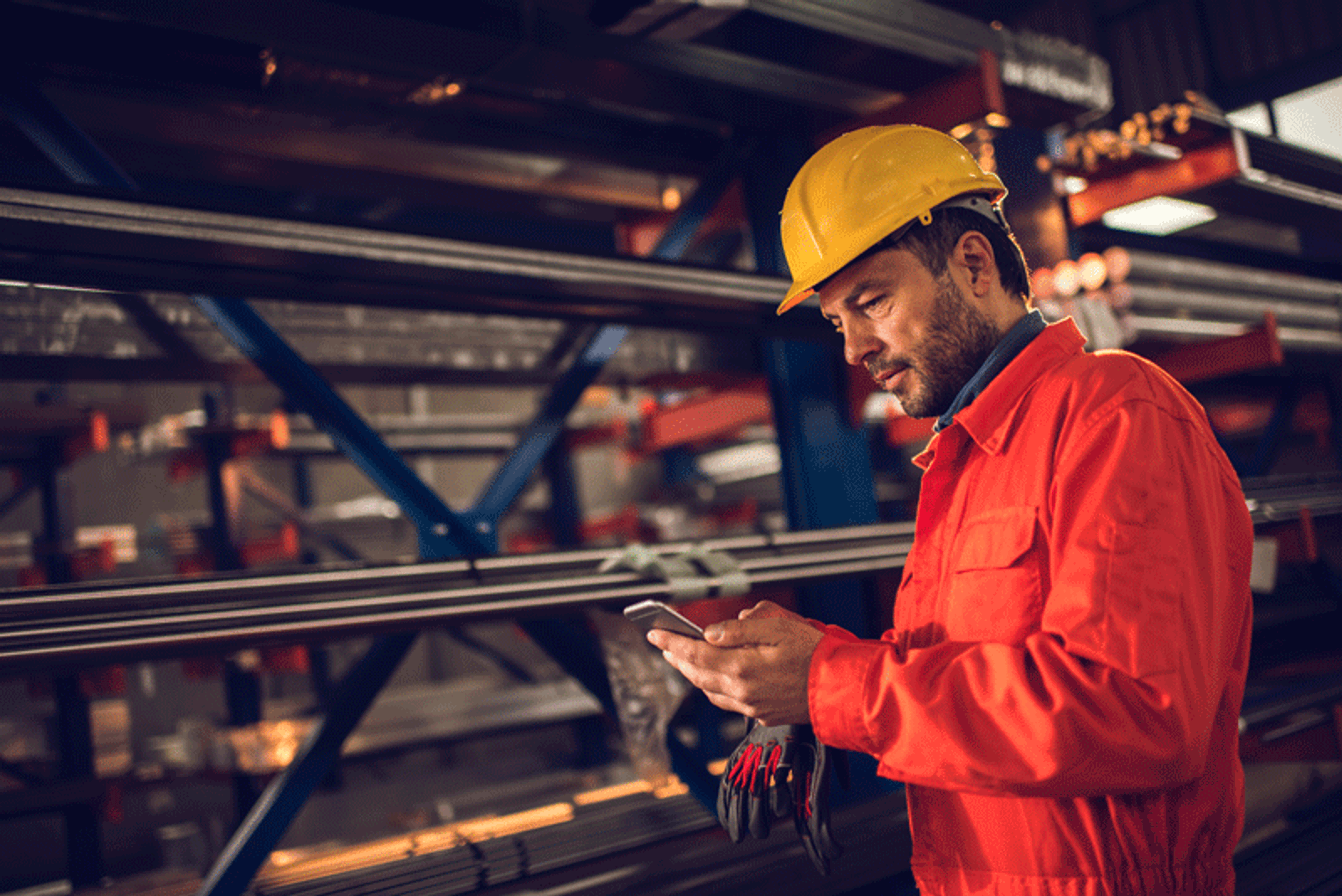 En industriarbeider i rød arbeidsdress og gul hjelm bruker en mobiltelefon foran hyller med metallbjelker.
