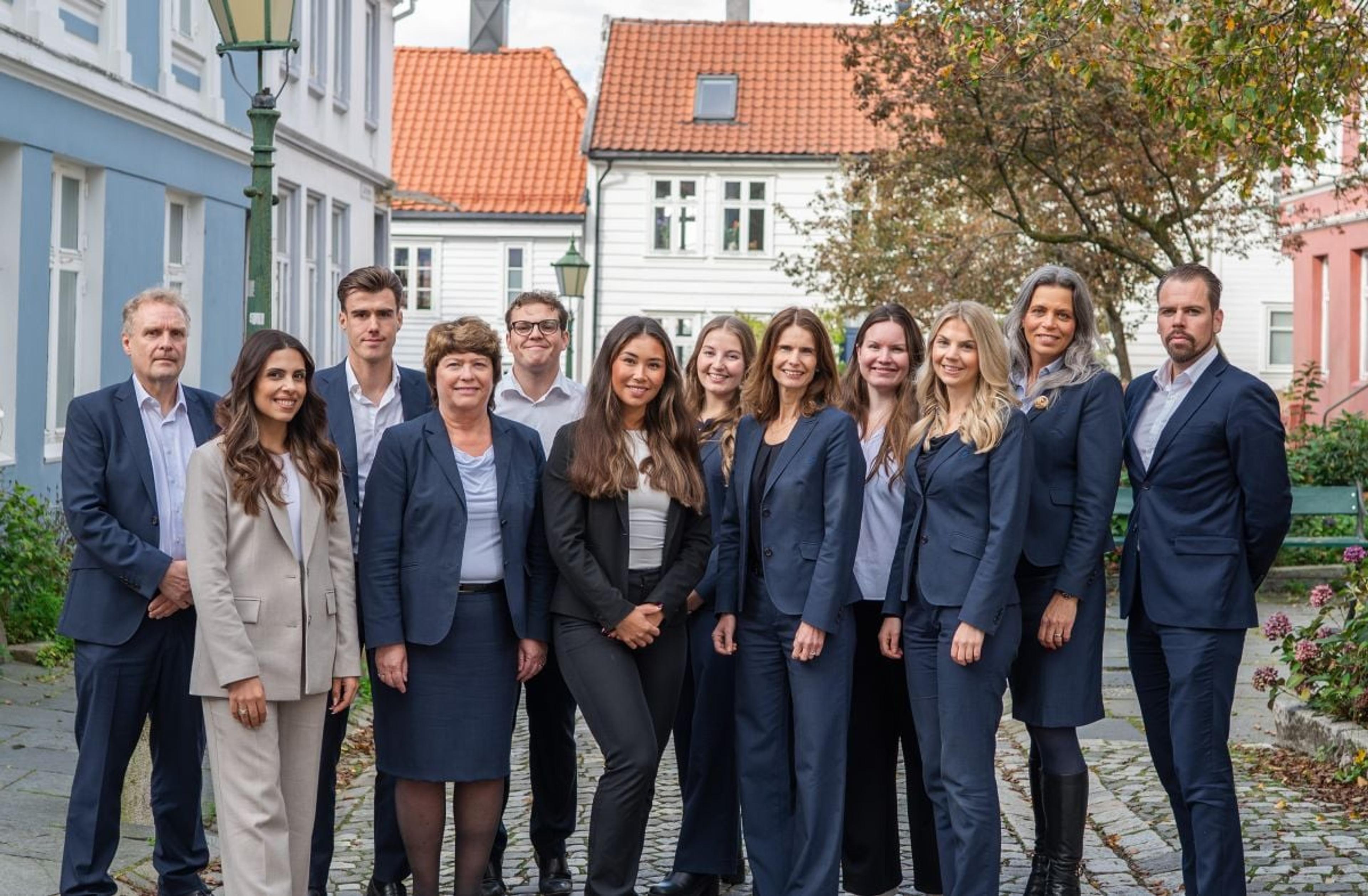 En gruppe på elleve kvinner og to menn i formelle dresser står samlet og smiler mot kameraet i en brosteinsgate med fargerike trehus i bakgrunnen. Bildet gir inntrykk av et profesjonelt team eller kollegium utendørs.