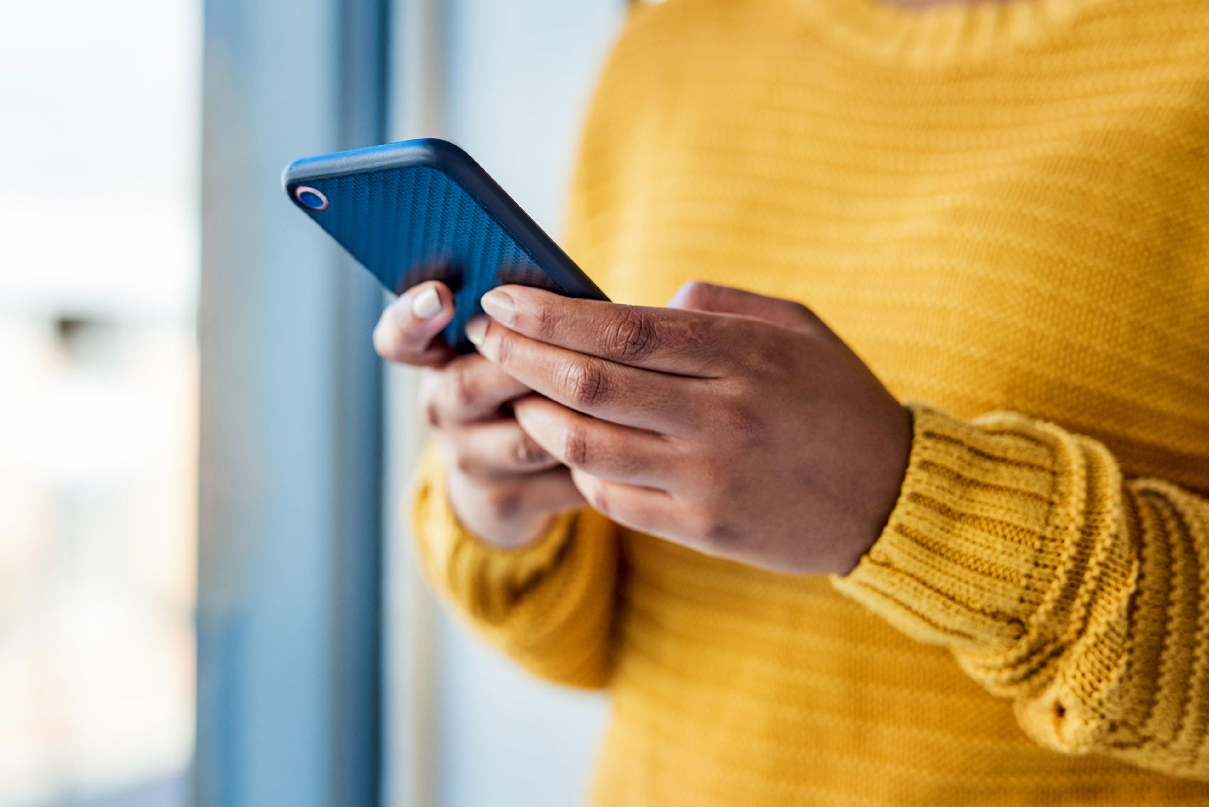 Nærbilde av en person i gul strikkegenser som holder og bruker en smarttelefon.