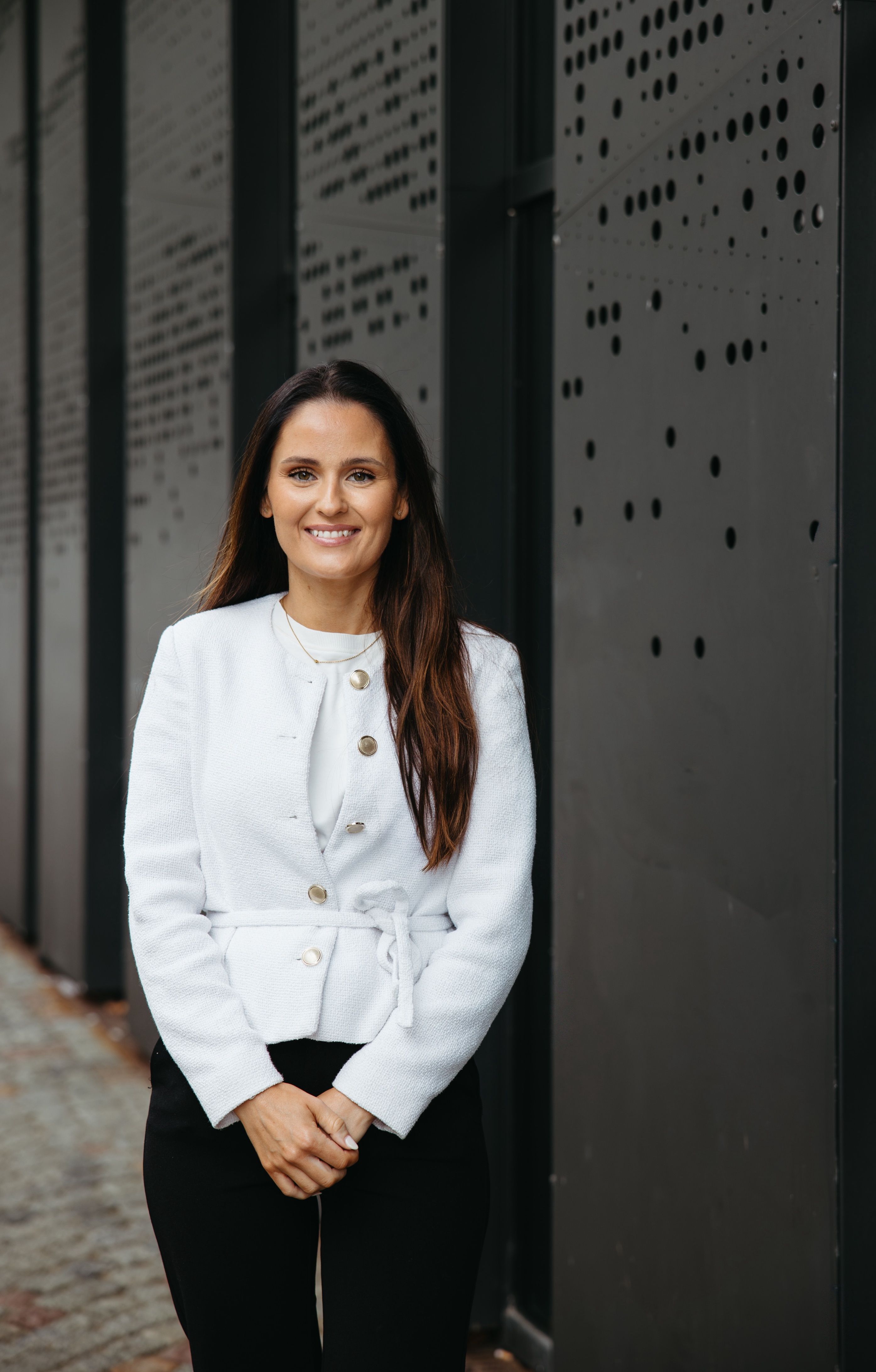 En smilende kvinne med langt, mørkt hår står foran en mørk, perforert fasade. Hun har på seg en hvit jakke med belte og svarte bukser.