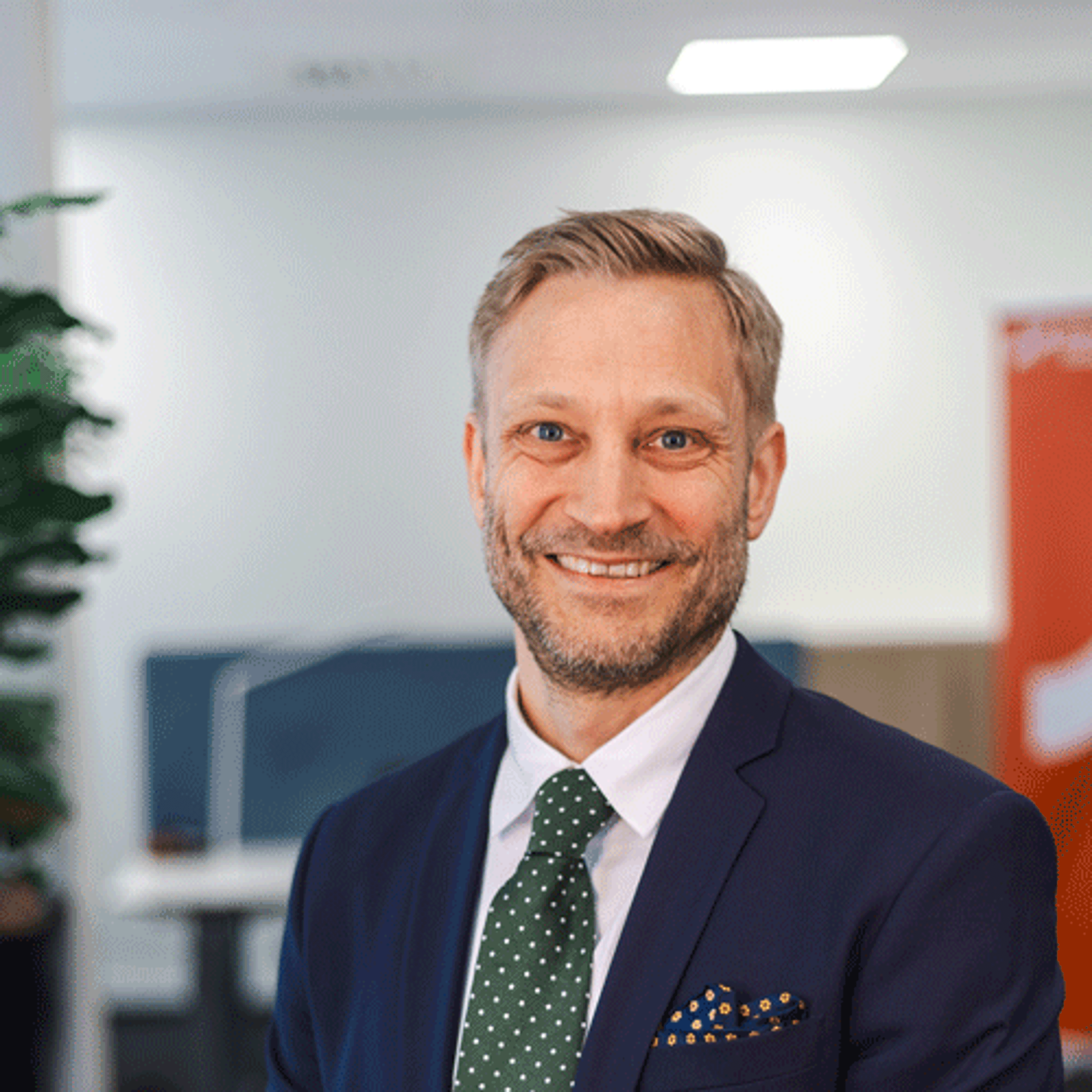 Smilende mann i dress og grønn slips står i et moderne kontormiljø med skrivebord og kontorskjermer i bakgrunnen.
