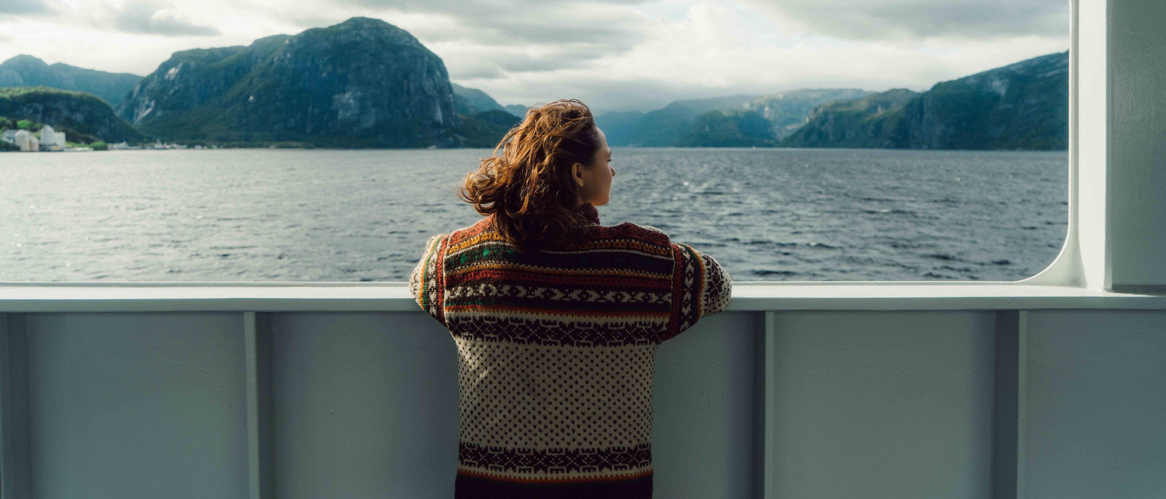 En person i mønstret strikkegenser står ved rekka på en båt og ser utover en fjord omgitt av bratte fjell under en overskyet himmel.