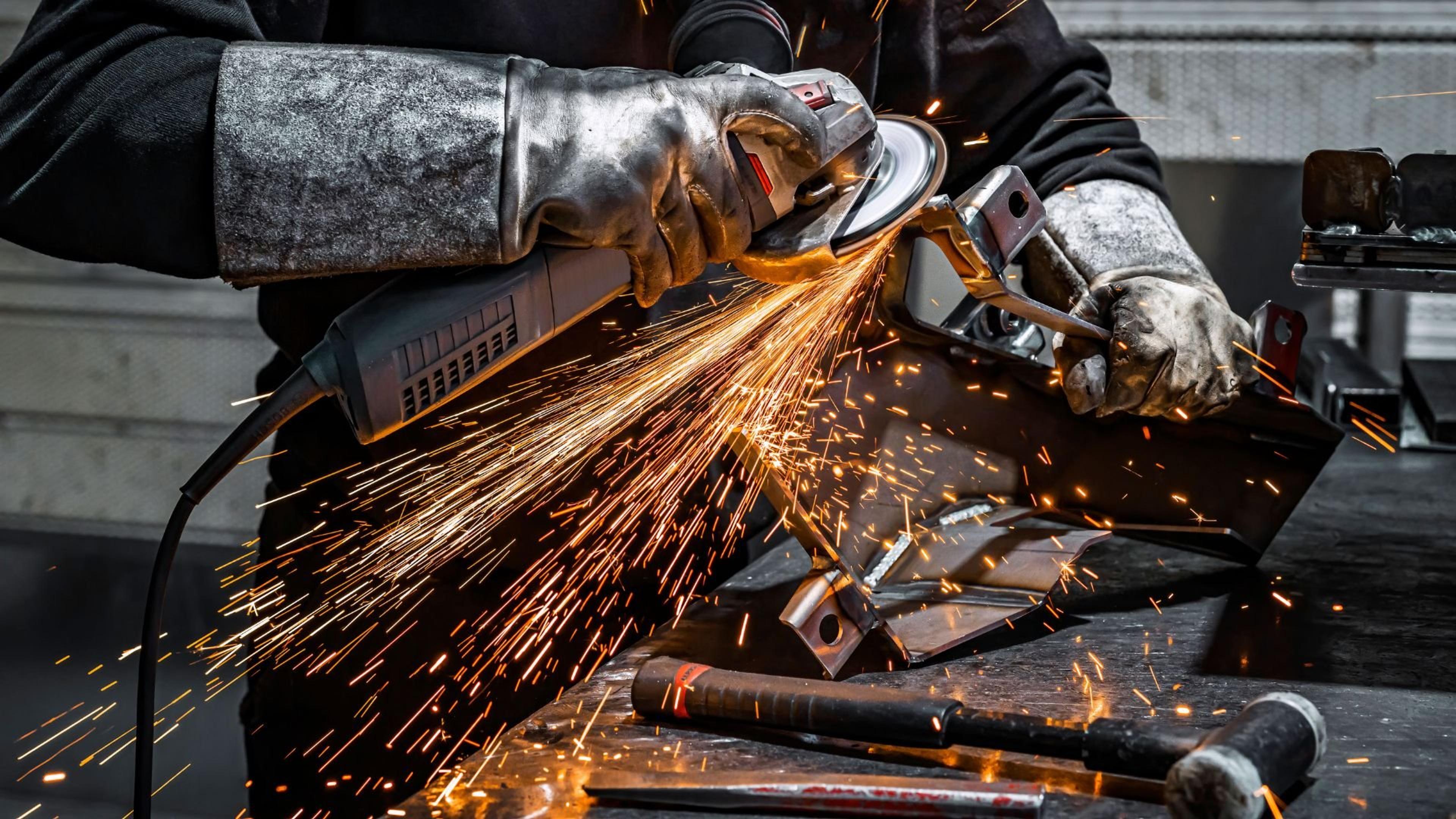 En arbeider med kraftige vernehansker bruker en vinkelsliper på en metalldel, mens en strøm av oransje gnister spruter over arbeidsbenken.