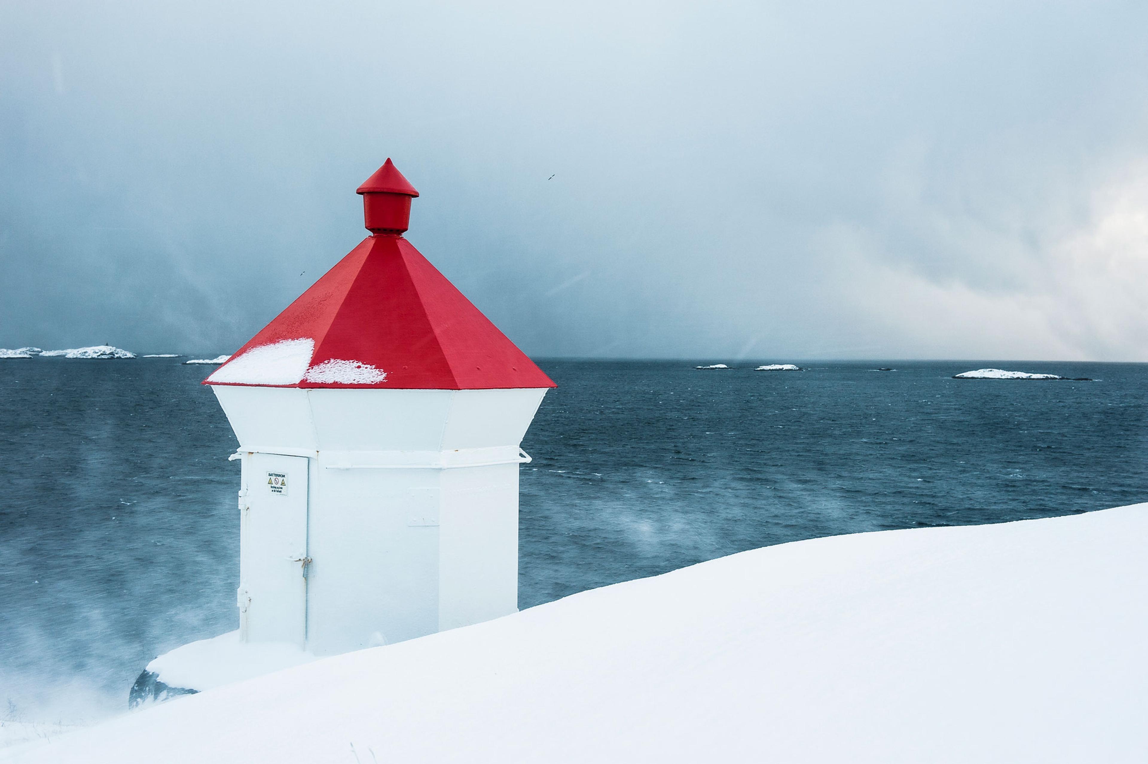 Et lite, hvitt fyr med rødt tak står i et snødekt kystlandskap ved et mørkt, urolig hav under en overskyet himmel.