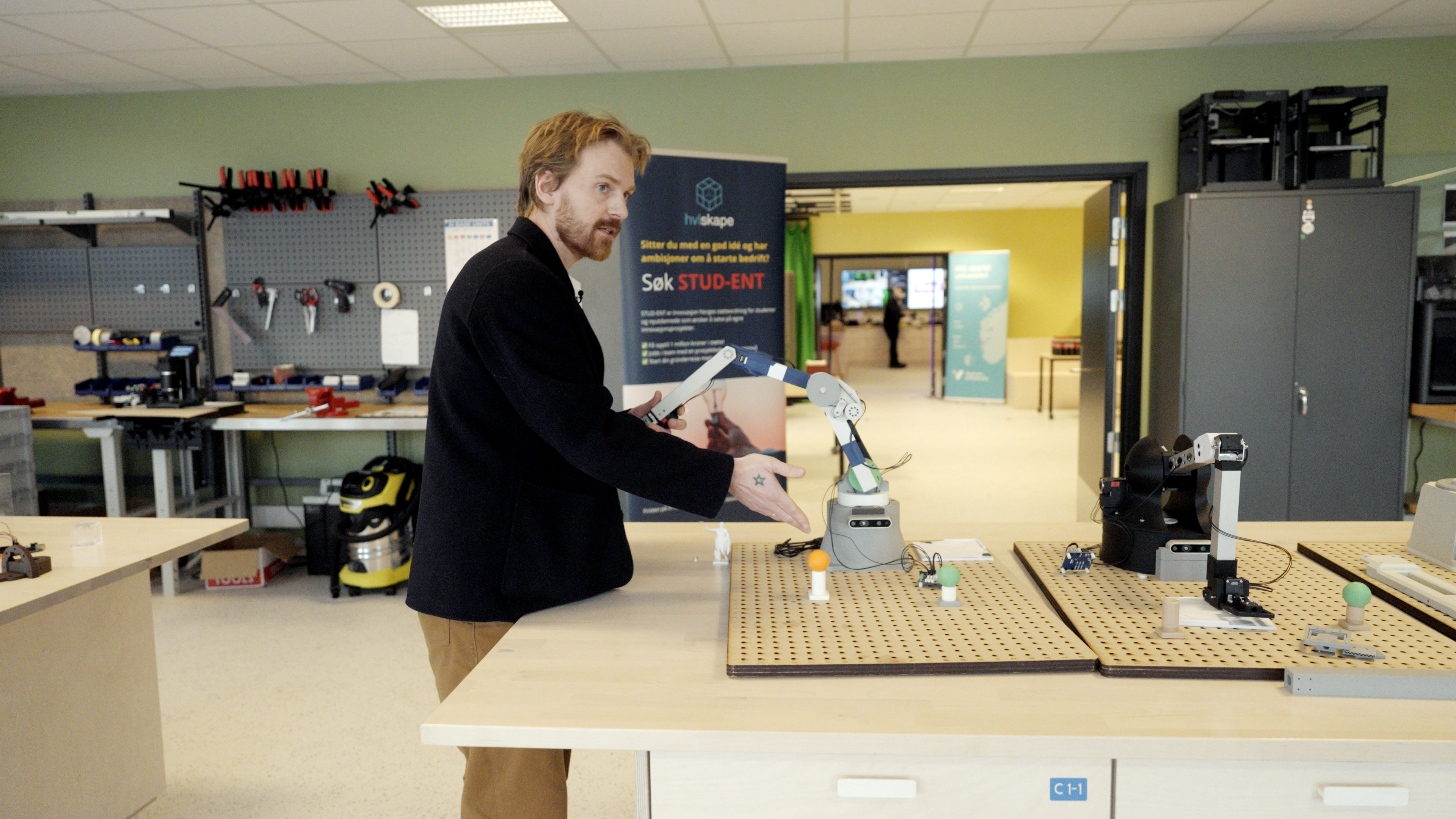 En mann demonstrerer små robotarmer på et arbeidsbord i et teknologiverksted. Verktøy og annet laboratorieutstyr er synlig i bakgrunnen.