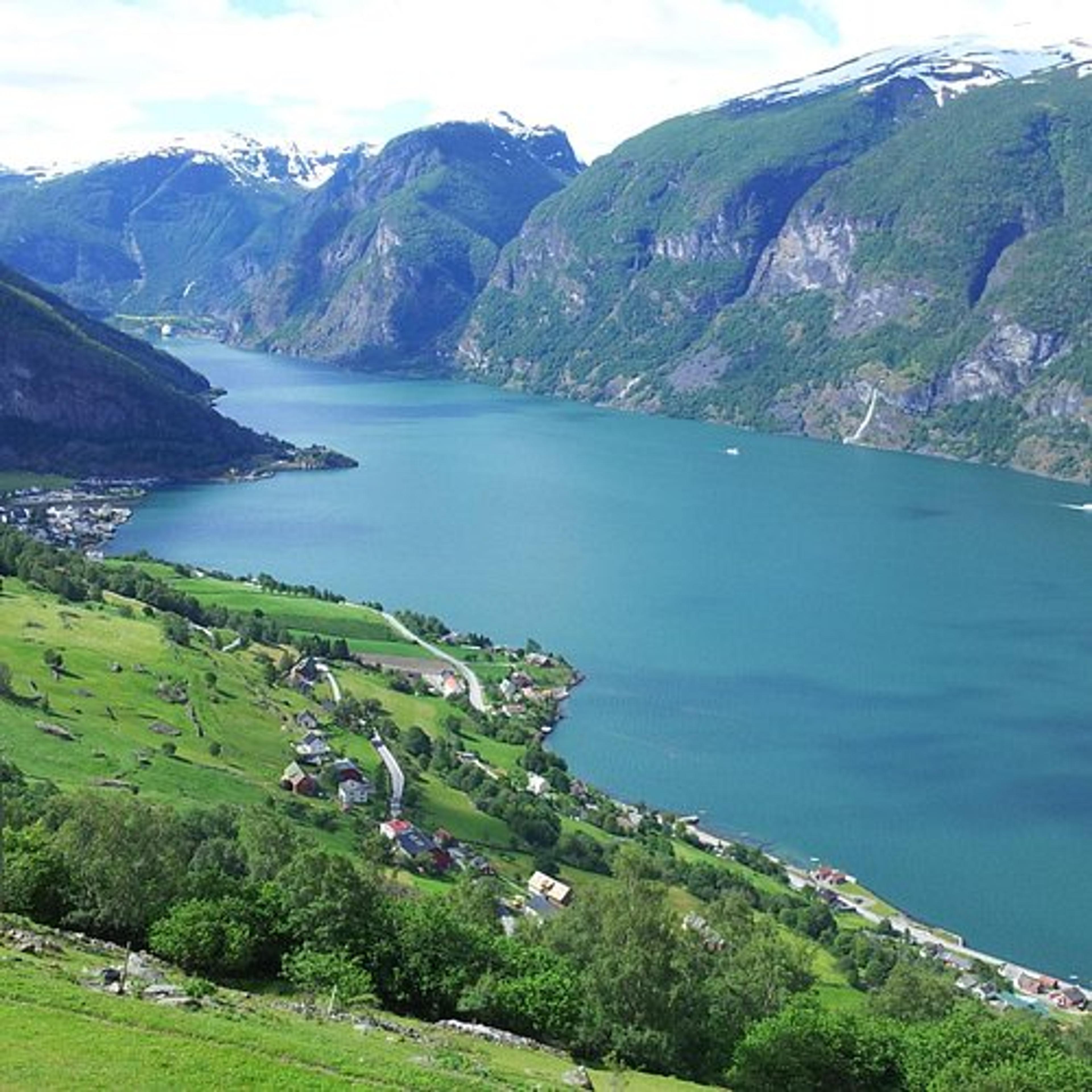 Utsikt over en dyp blå fjord omgitt av grønne, bratte fjellsider med snødekte topper, med en liten bygd og gårder langs den grønne dalen ned mot vannet.