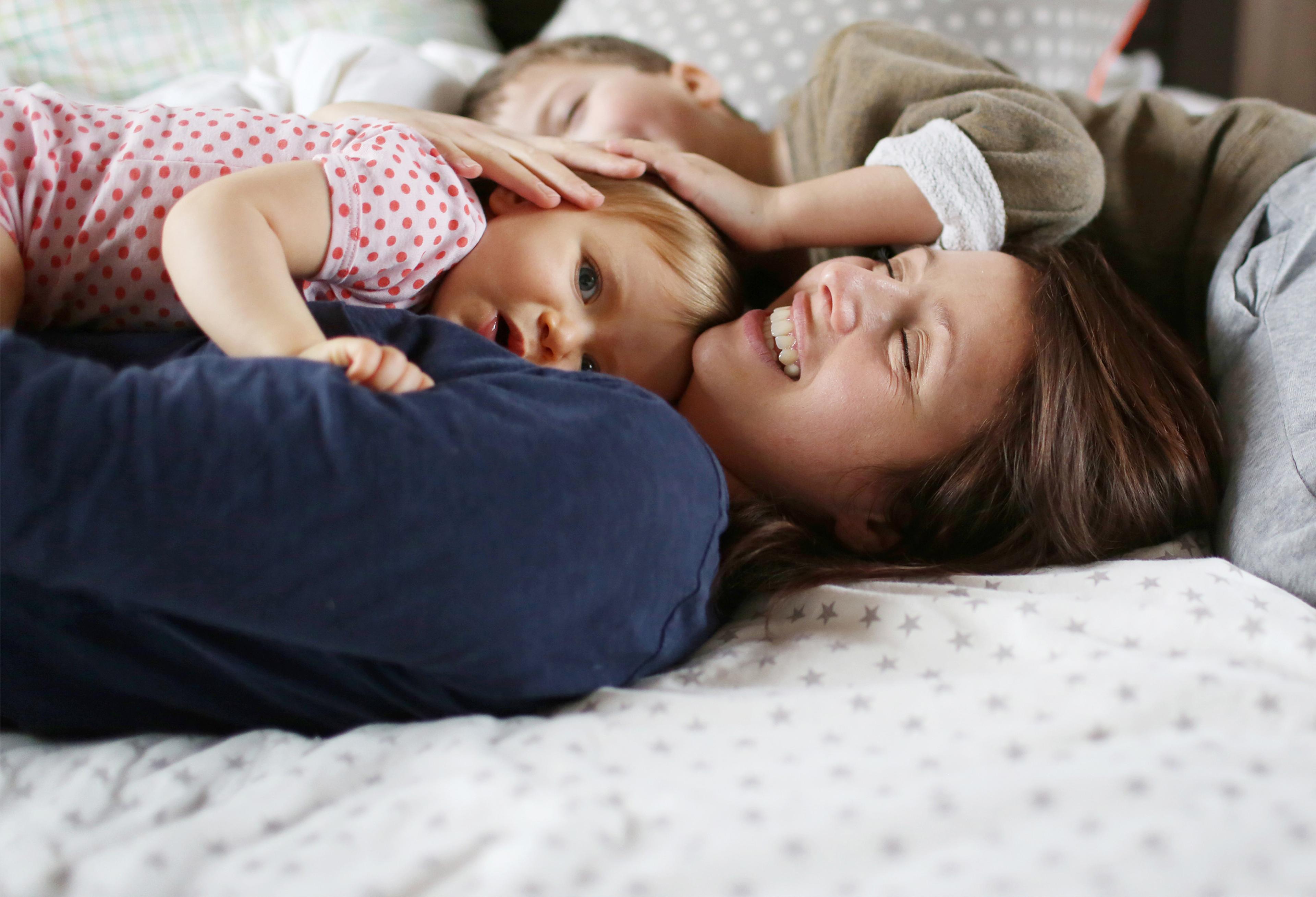 En smilende mor ligger i sengen og koser med en baby, mens et eldre barn ligger tett inntil dem.