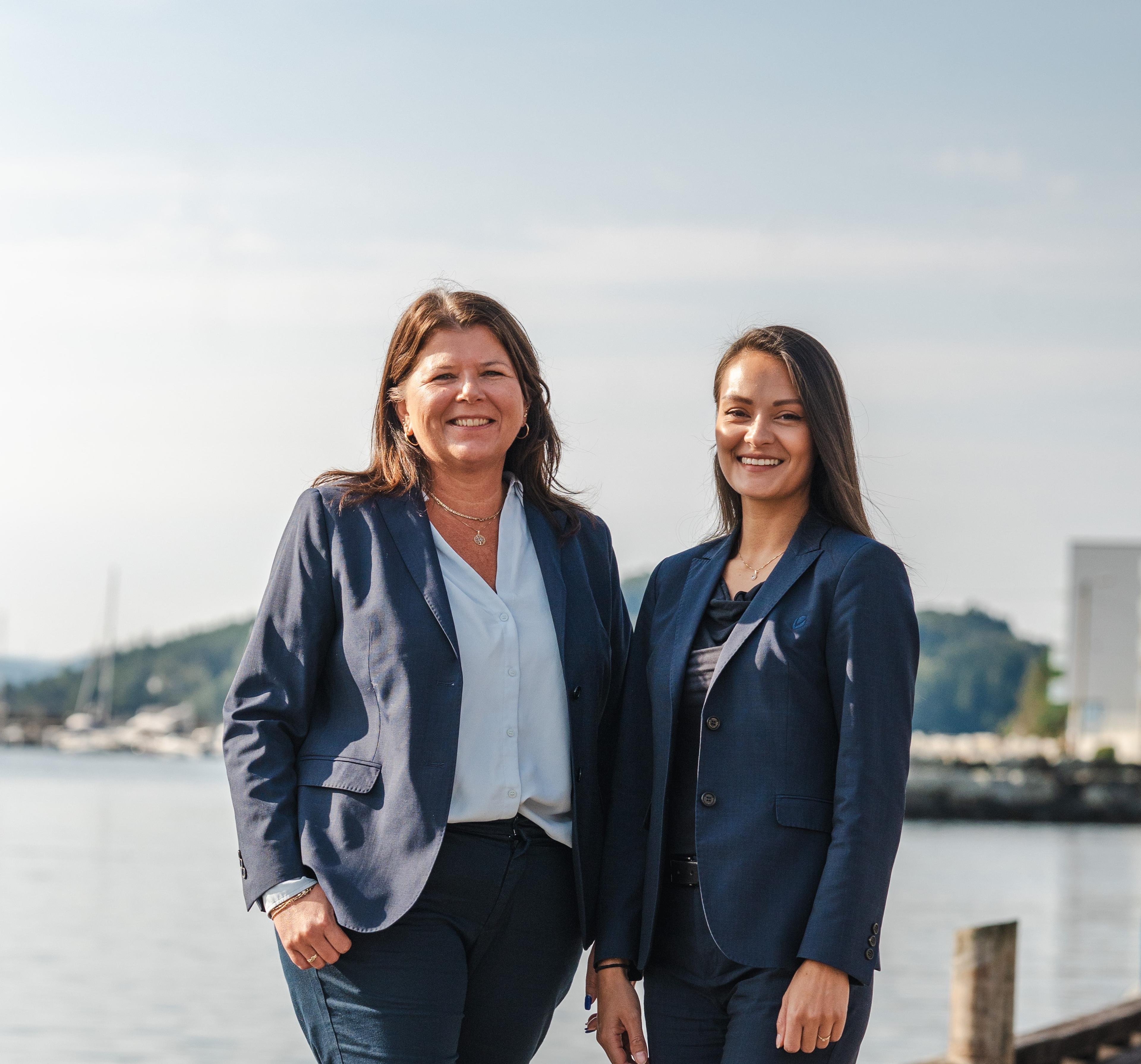 To smilende kvinner i mørkeblå dresser står sammen ved vannet, med en kystlinje i bakgrunnen.