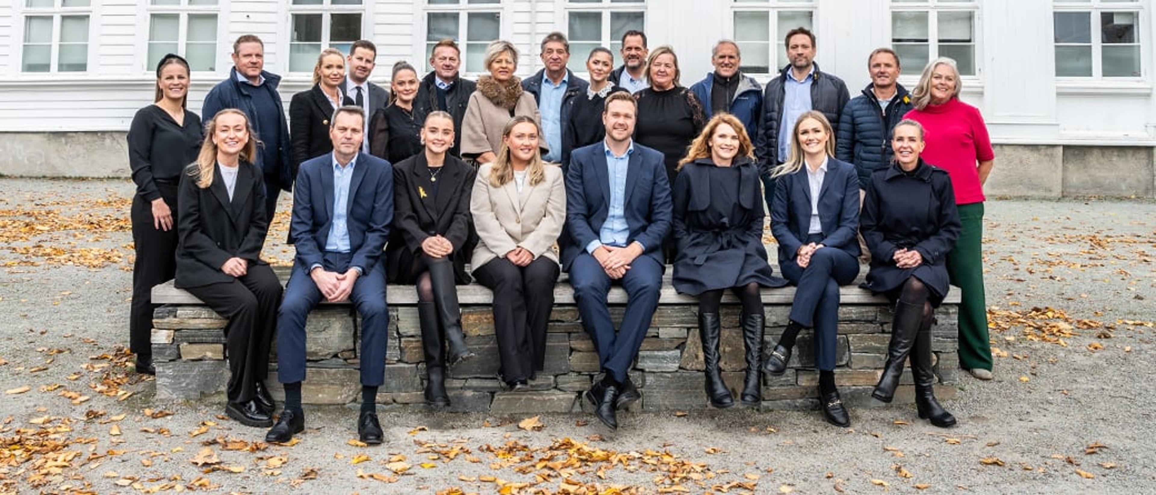 En større gruppe voksne personer poserer smilende for et gruppebilde utendørs foran en hvit bygning, mange sittende på en lav steinmur og noen stående bak. Det ligger gule høstblader på bakken foran dem.