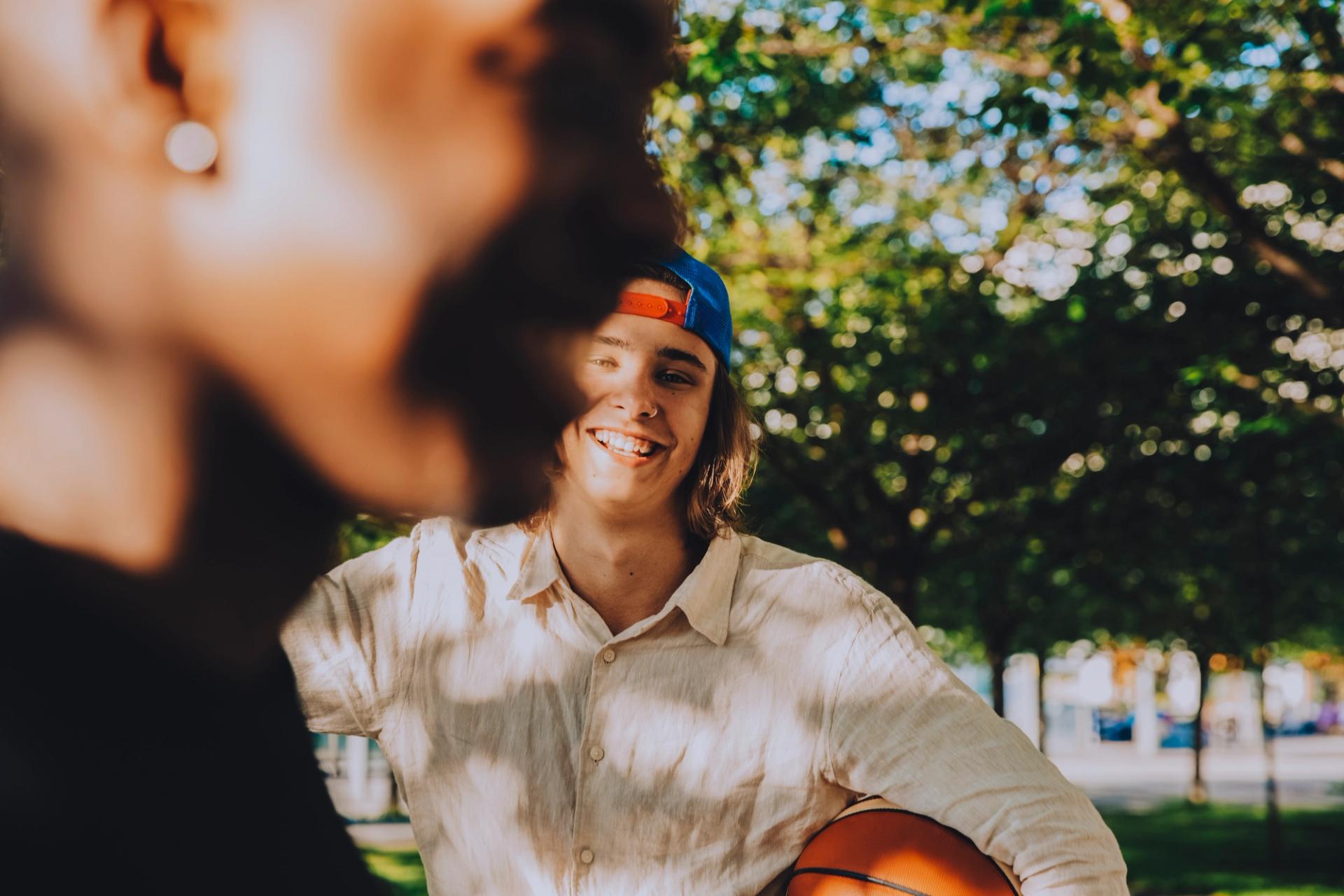 En smilende ung person med bakvendt caps holder en basketball i en solfylt park, mens en annen person er uskarp i forgrunnen.