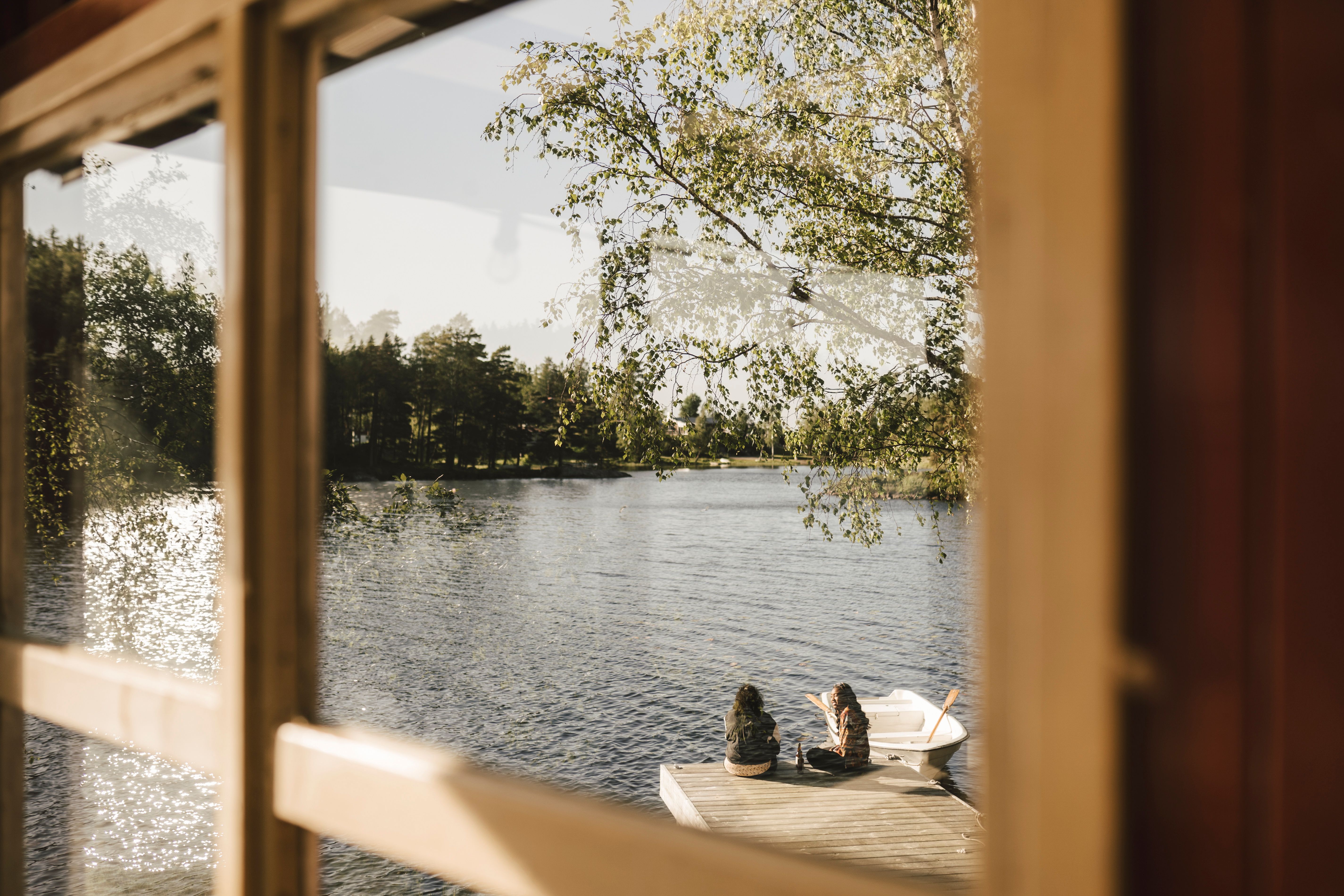 Utsikt gjennom et vindu mot en solfylt innsjø, der to personer sitter på en brygge ved siden av en liten robåt.