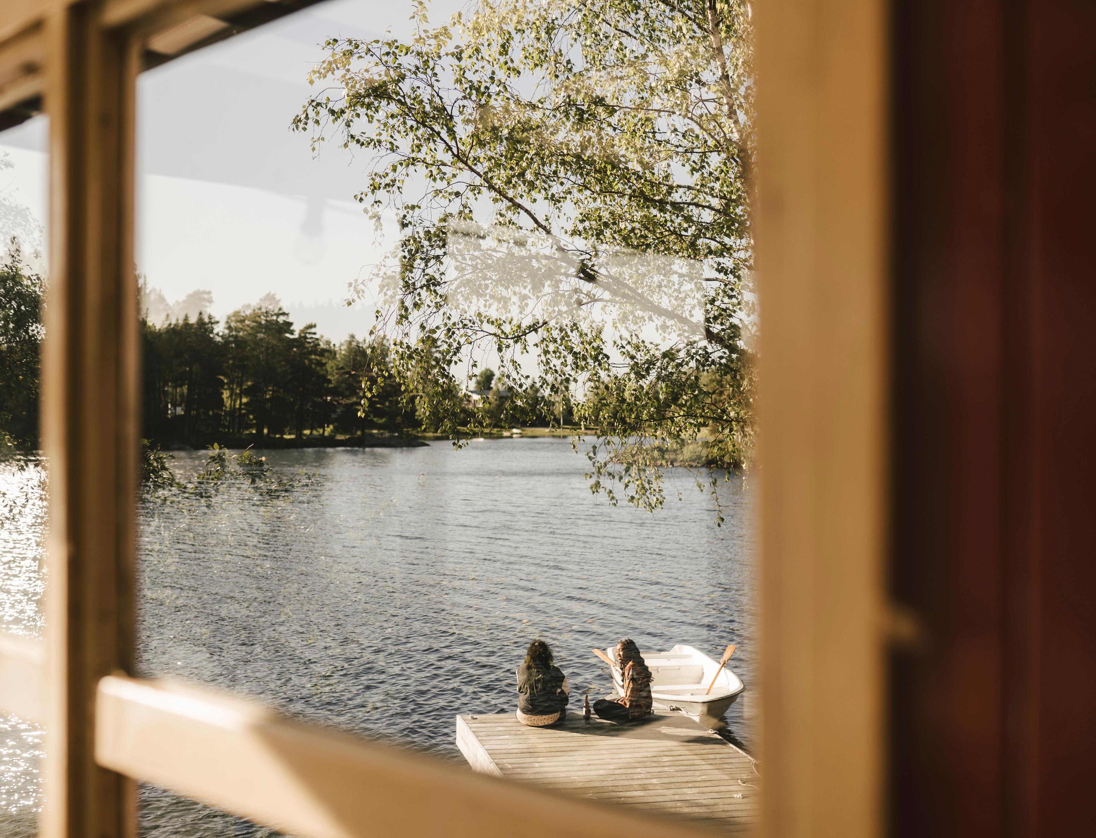 Utsikt gjennom et vindu mot en solfylt innsjø, der to personer sitter på en brygge ved siden av en liten robåt.