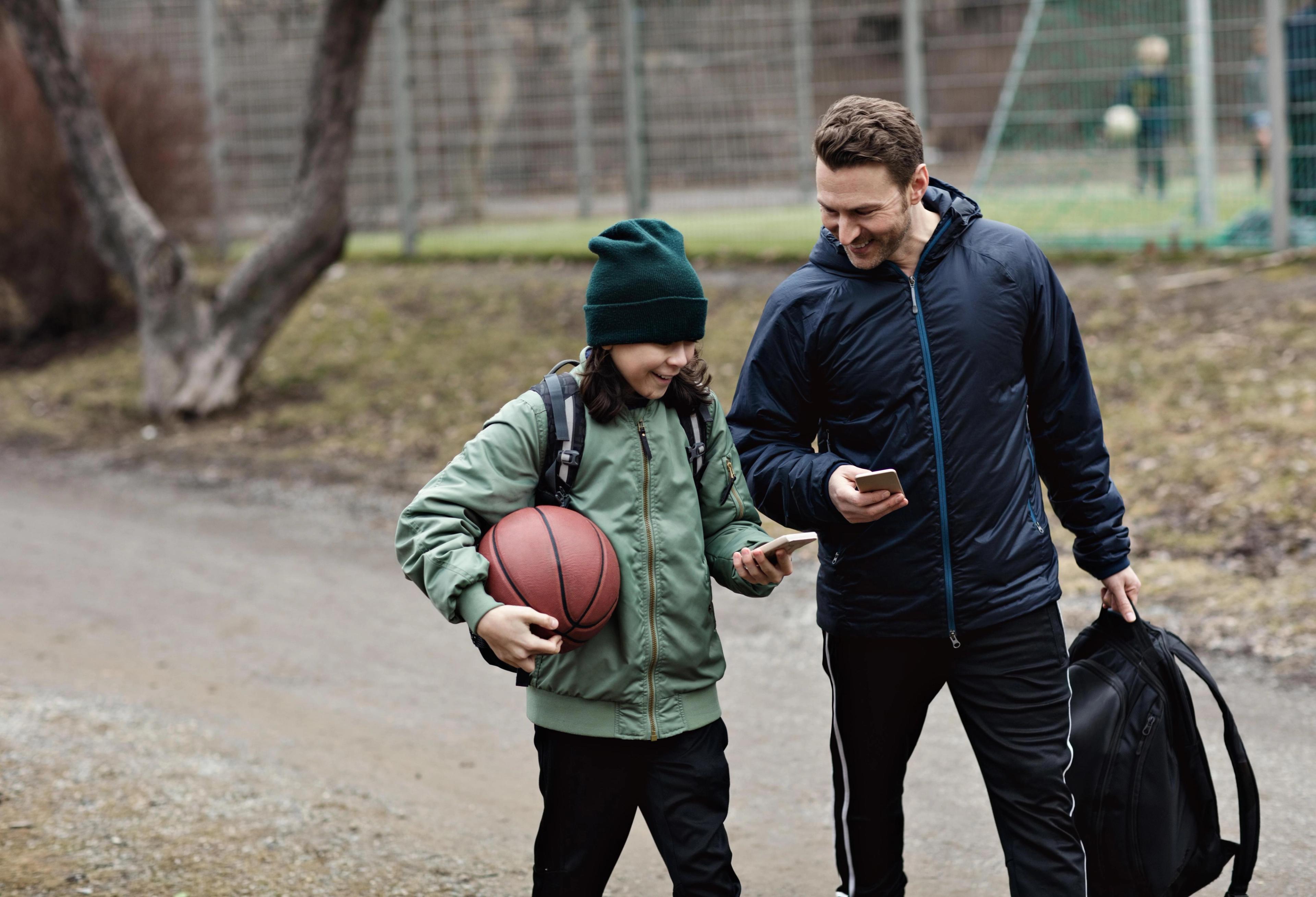 En gutt med basketball og en mann med treningsbag går sammen ved en inngjerdet idrettsbane mens de ser på mobiltelefonene sine.