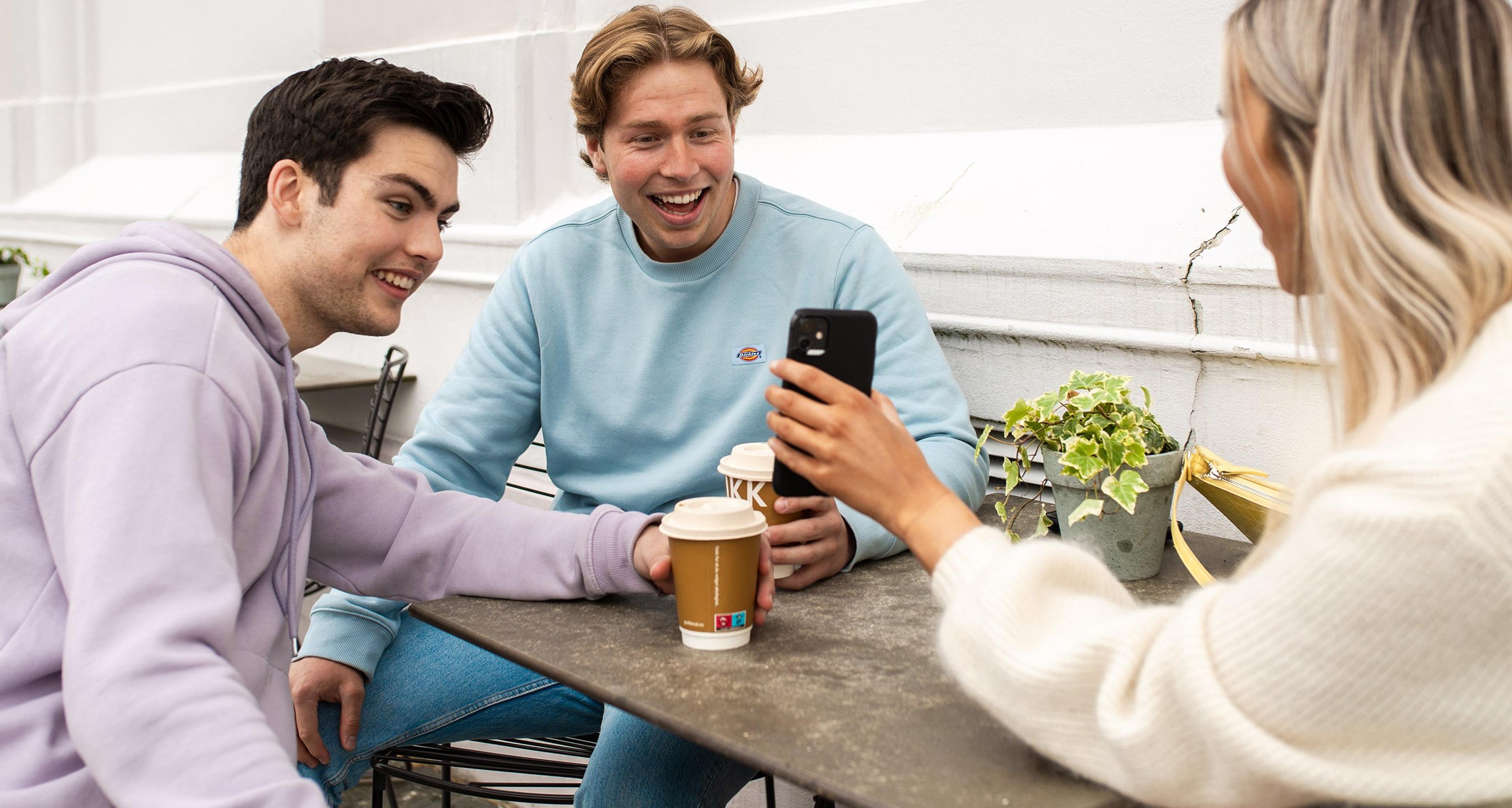 To personer sitter ved et bord med kaffekopper og ser på en mobiltelefon som viser en app med økonomisk oversikt.