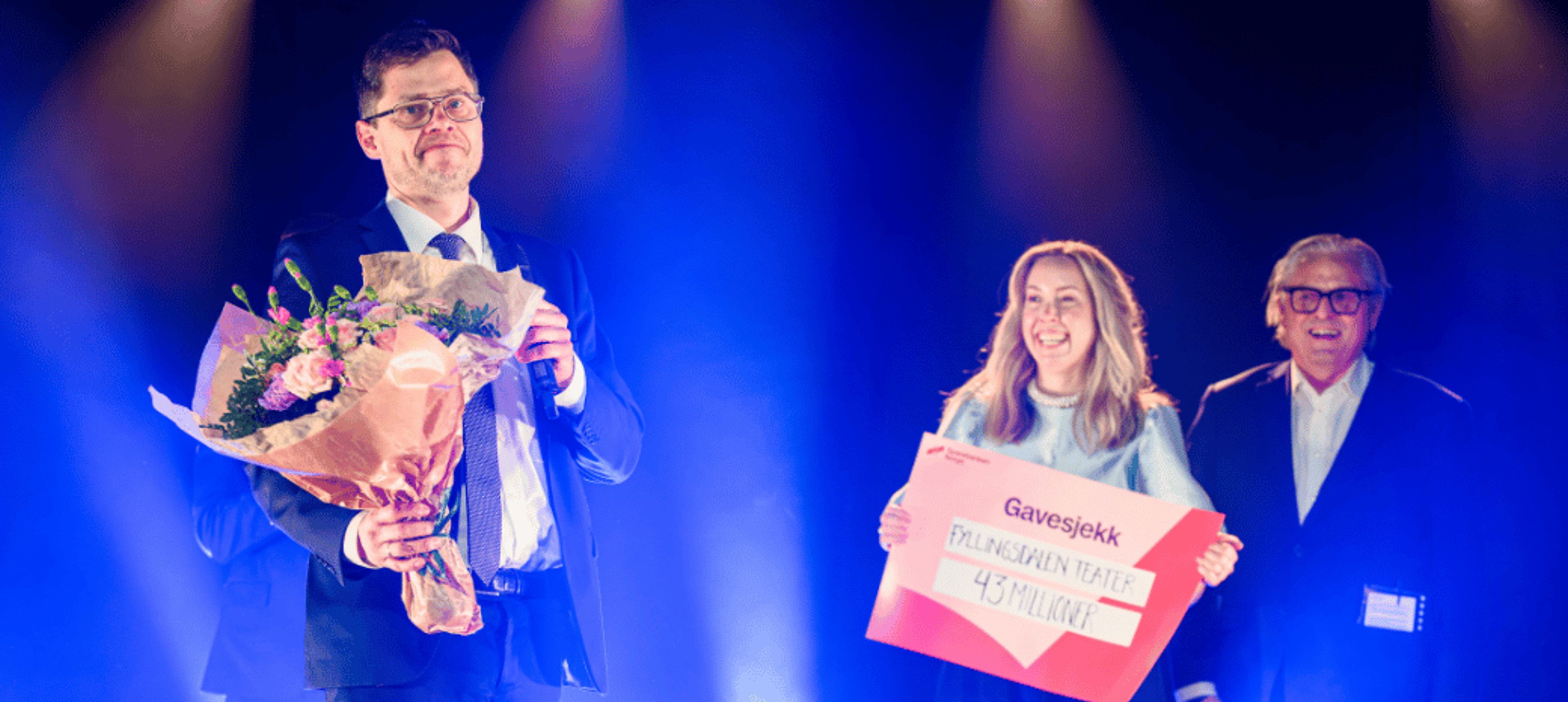 En mann i dress holder en stor blomsterbukett på en blåbelyst scene, mens en smilende kvinne viser frem en stor gavesjekk på 43 millioner kroner ved siden av en annen mann.
