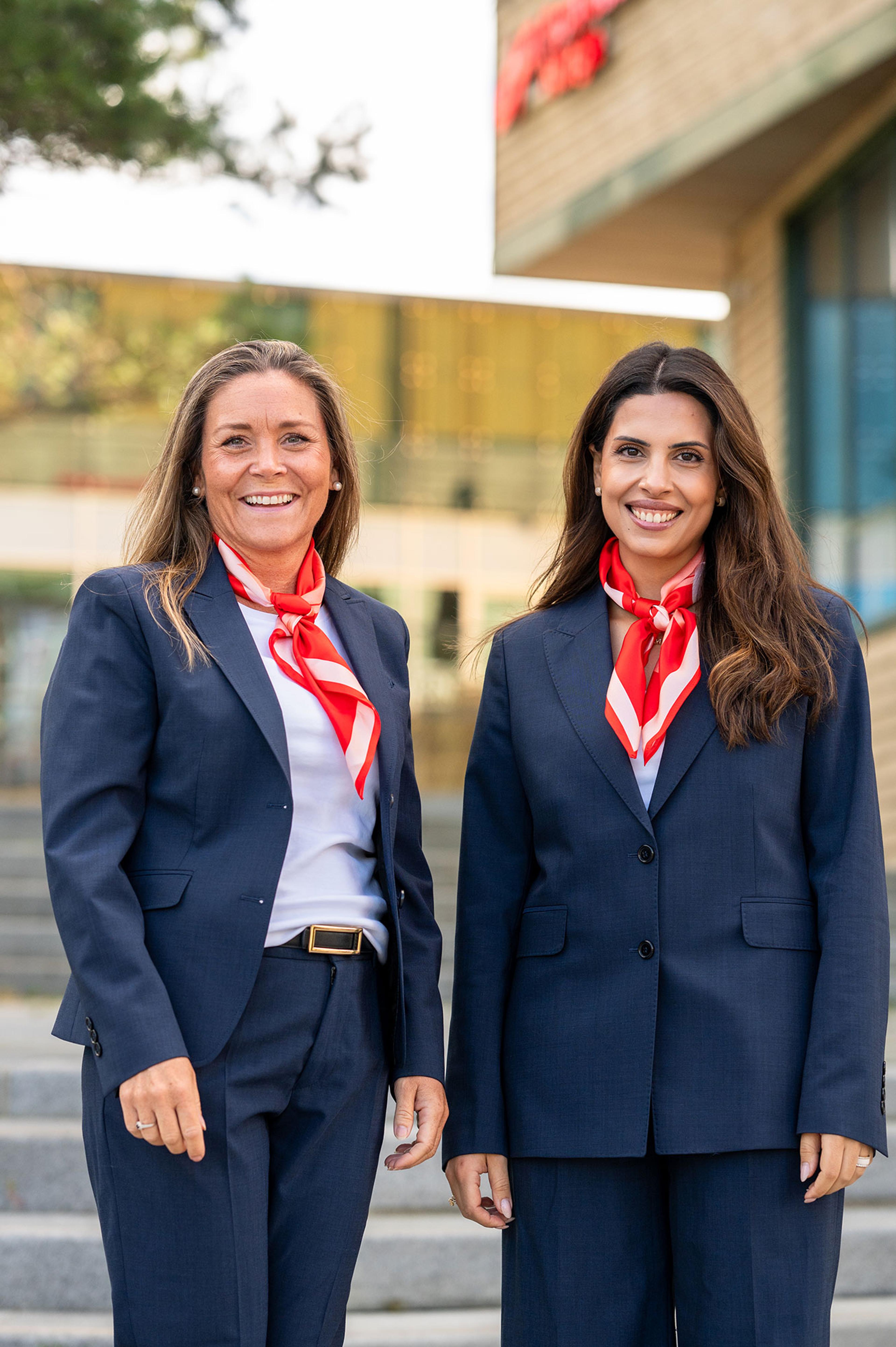 To smilende kvinner i mørkeblå uniformsdresser og røde halstørklær står utendørs foran en moderne bygning.