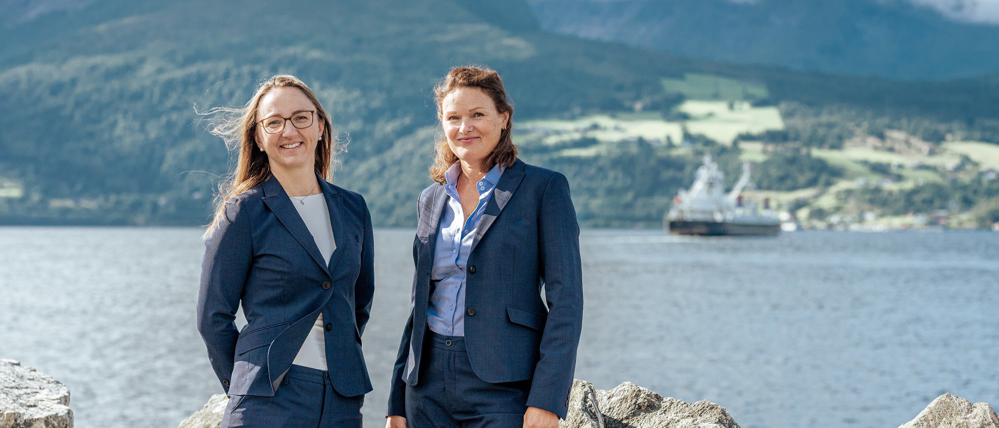 To kvinner i mørke blå dresser står og smiler ved en steinmur foran en fjord, med en ferge og grønne åser i bakgrunnen.