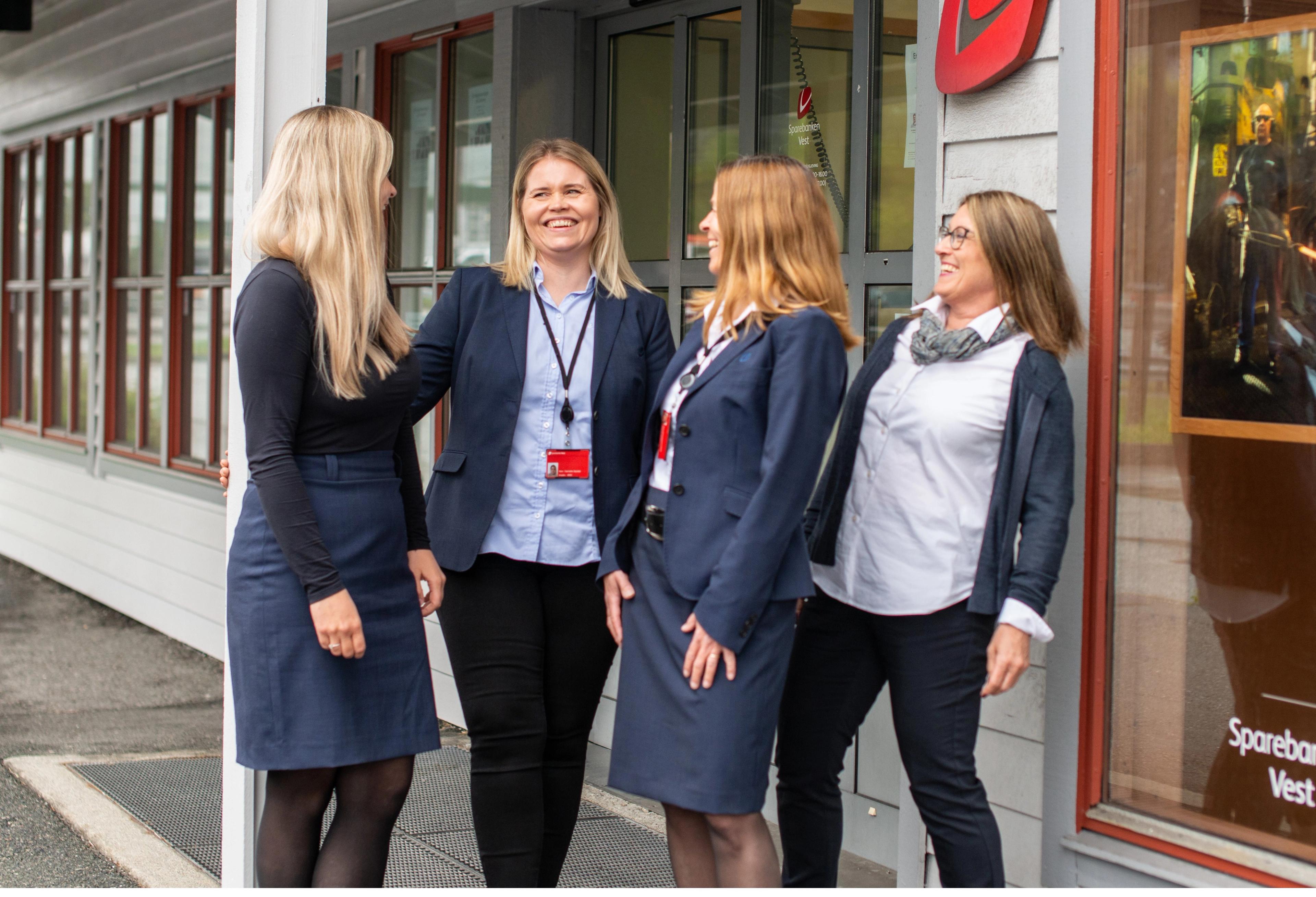 Fire kvinnelige bankansatte i dress og navneskilt står utenfor en bankfilial og smiler til hverandre. De ser avslappede og kollegiale ut foran inngangspartiet.