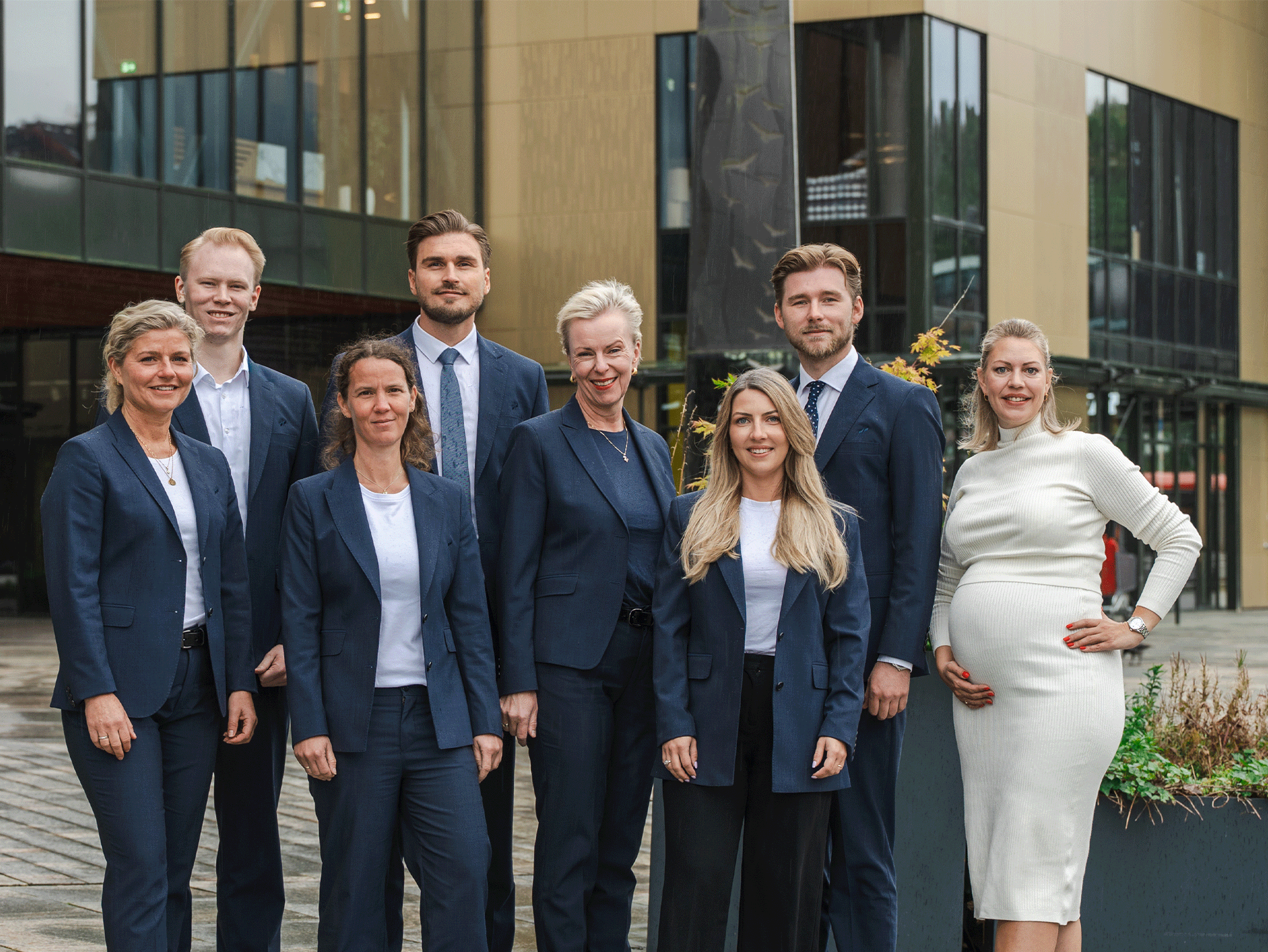 Åtte profesjonelt kledde personer står samlet foran en moderne kontorbygning. Gruppen består av fem kvinner og tre menn, og en av kvinnene er gravid.