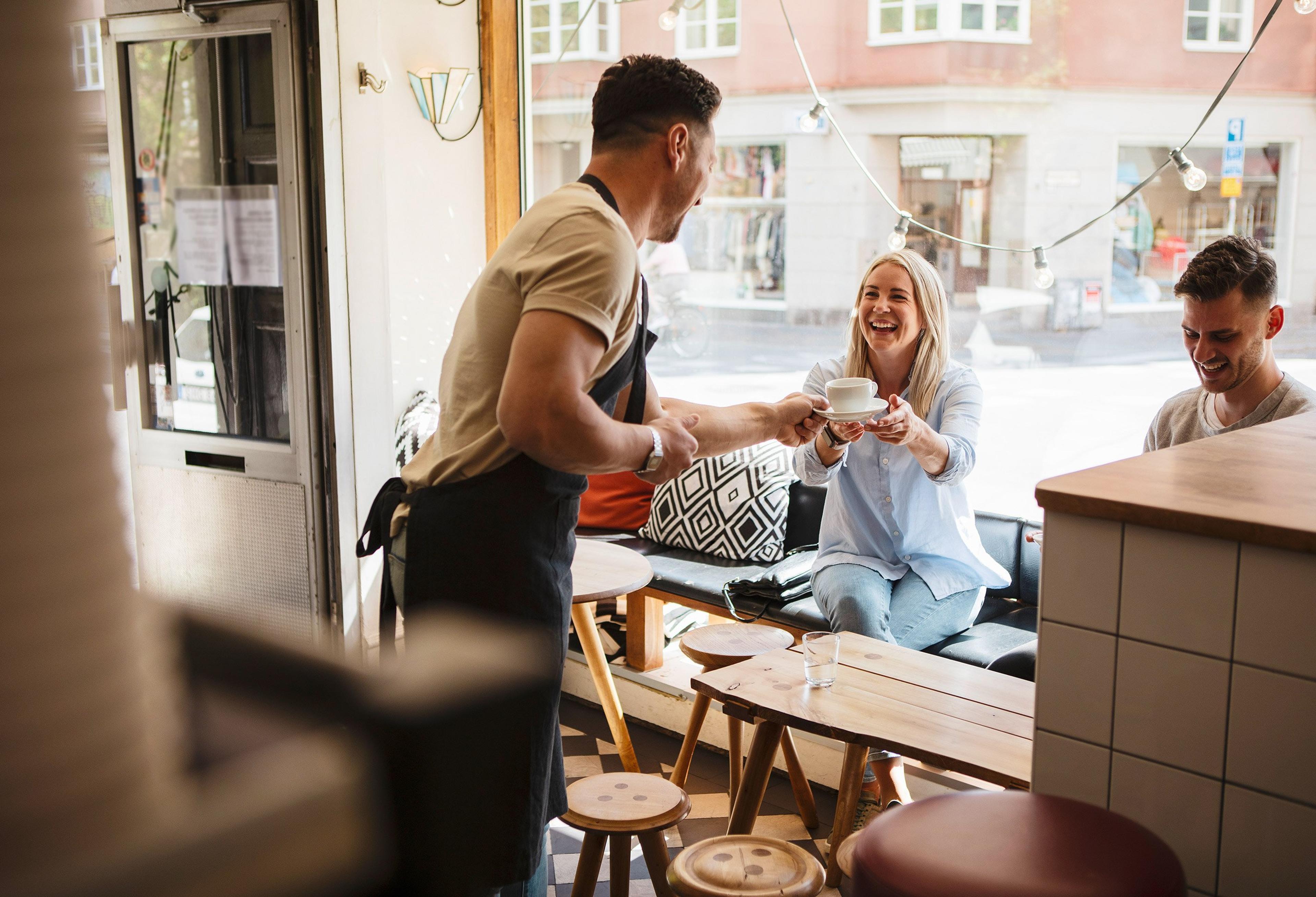 En smilende servitør i forkle rekker en kopp kaffe til en kvinne som sitter ved et bord i en lys og hyggelig kafé, mens en mann ved siden av henne ser ned og smiler.