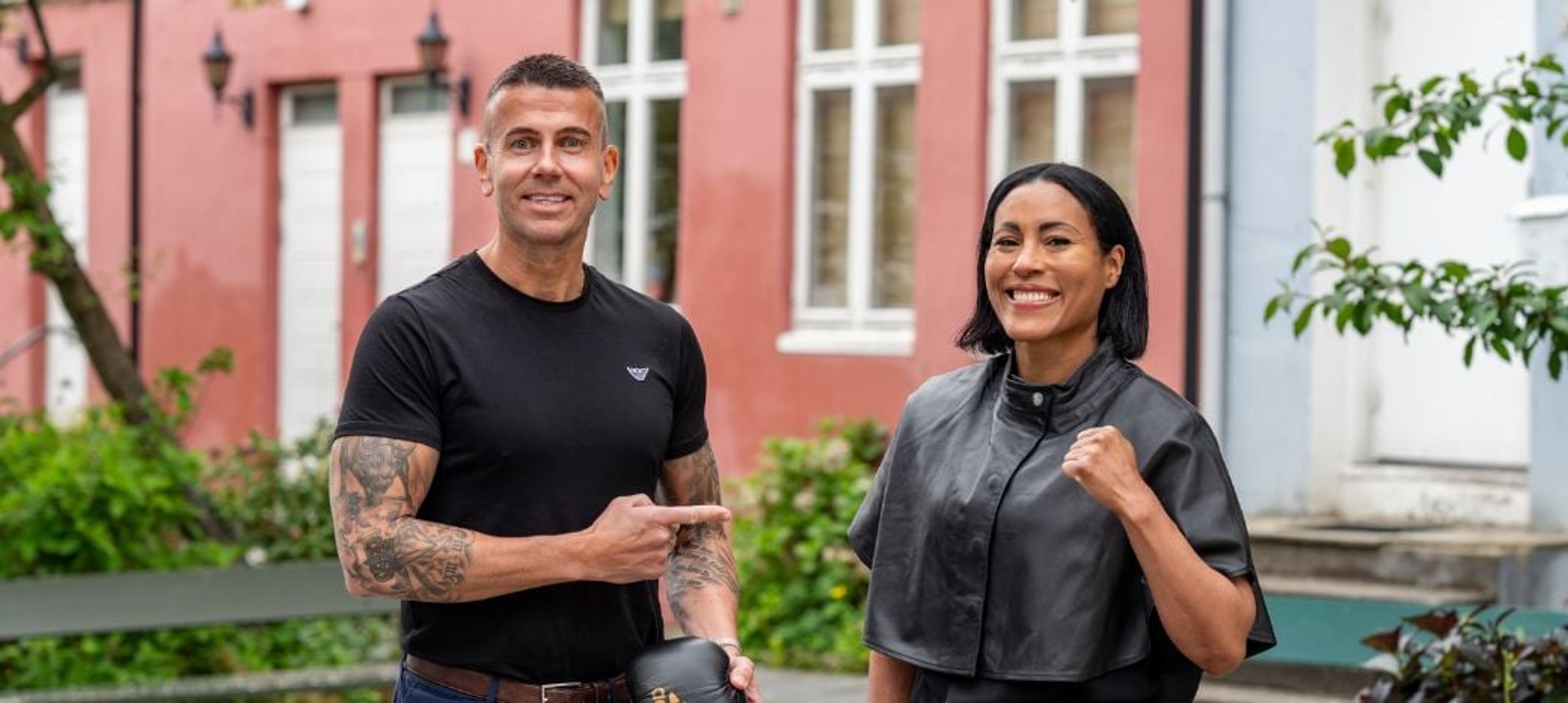 En smilende mann i svart T-skjorte peker mot en smilende kvinne i svart jakke, mens de står utendørs foran fargerike bygninger.