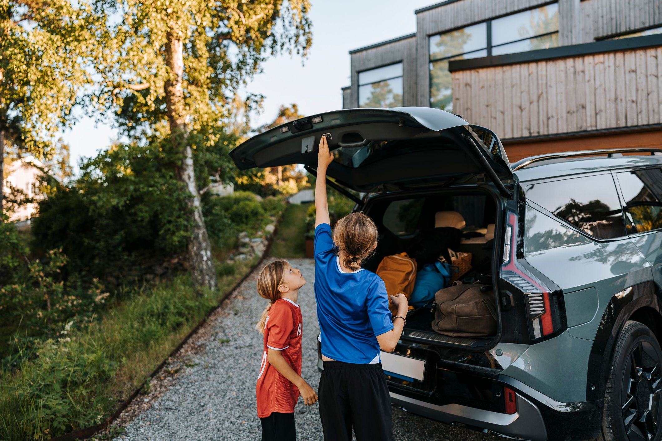 En kvinne lukker bakluken på en fullpakket SUV mens et barn står ved siden av i oppkjørselen foran et hus.