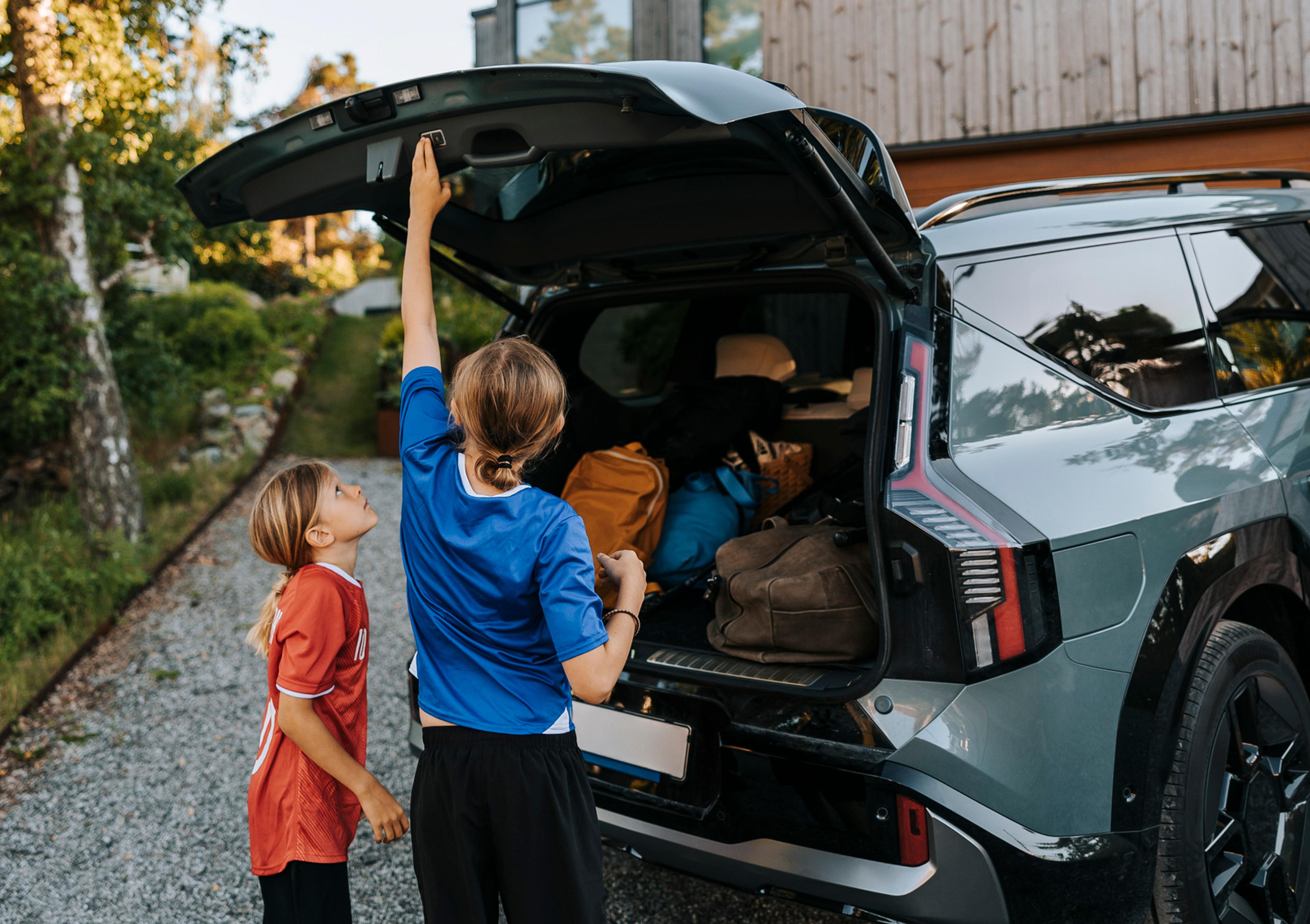 En kvinne lukker bakluken på en fullpakket SUV mens et barn står ved siden av i oppkjørselen foran et hus.