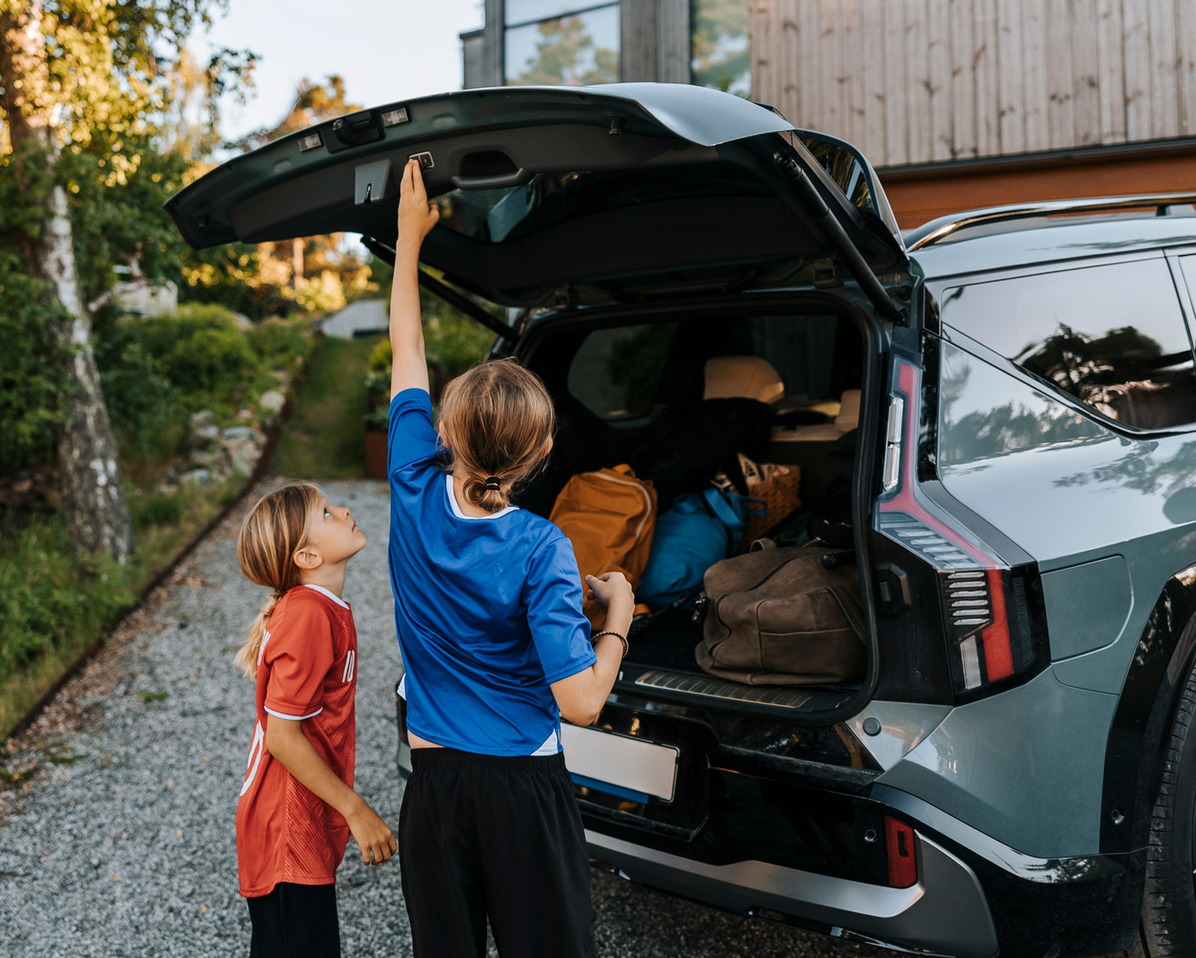 Et barn lukker bakluken på en fullpakket SUV mens et barn står ved siden av i oppkjørselen foran et hus.