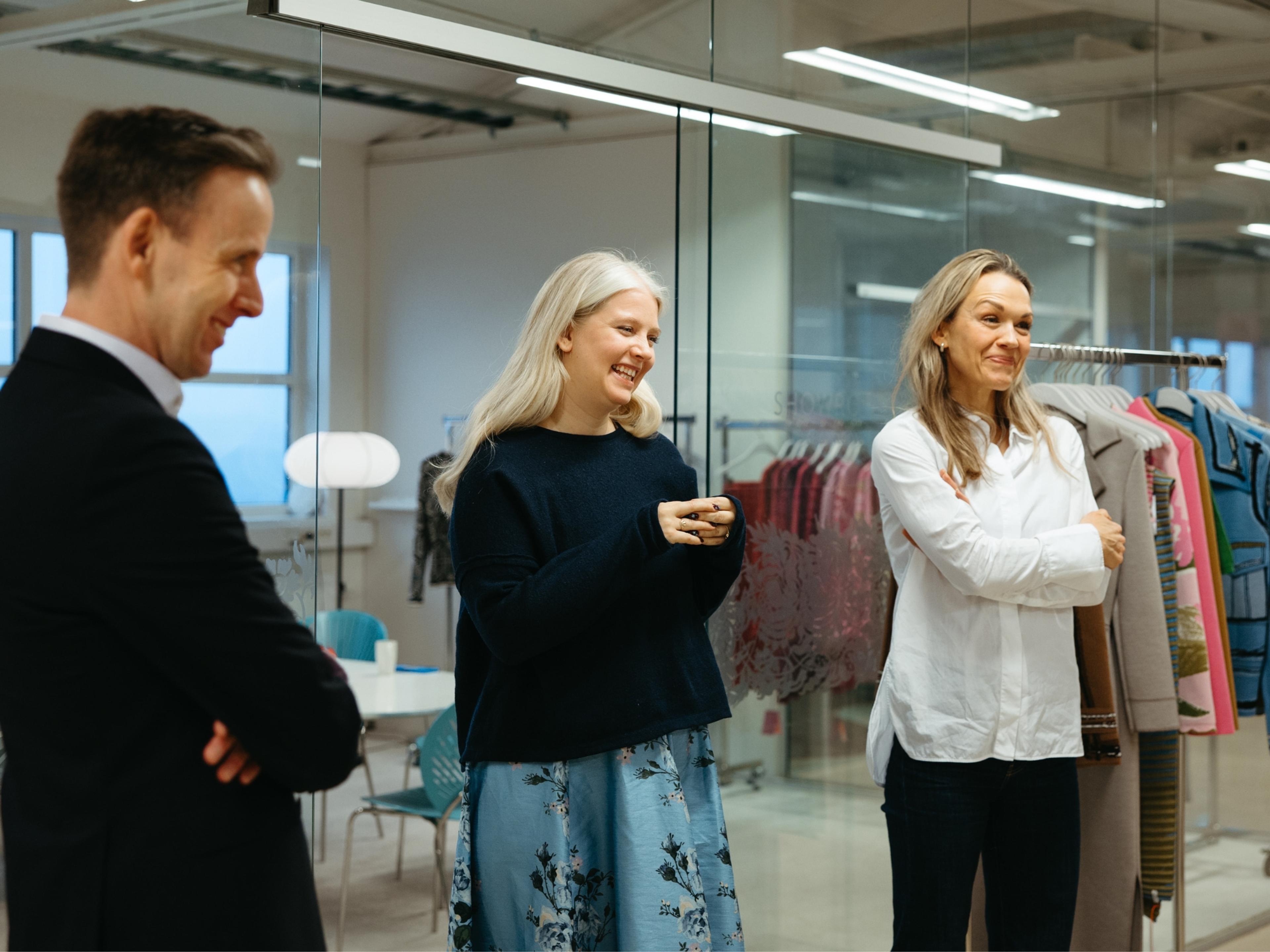 Tre personer står smilende sammen i et moderne kontor med glassvegger. I bakgrunnen henger fargerike klær på et stativ.