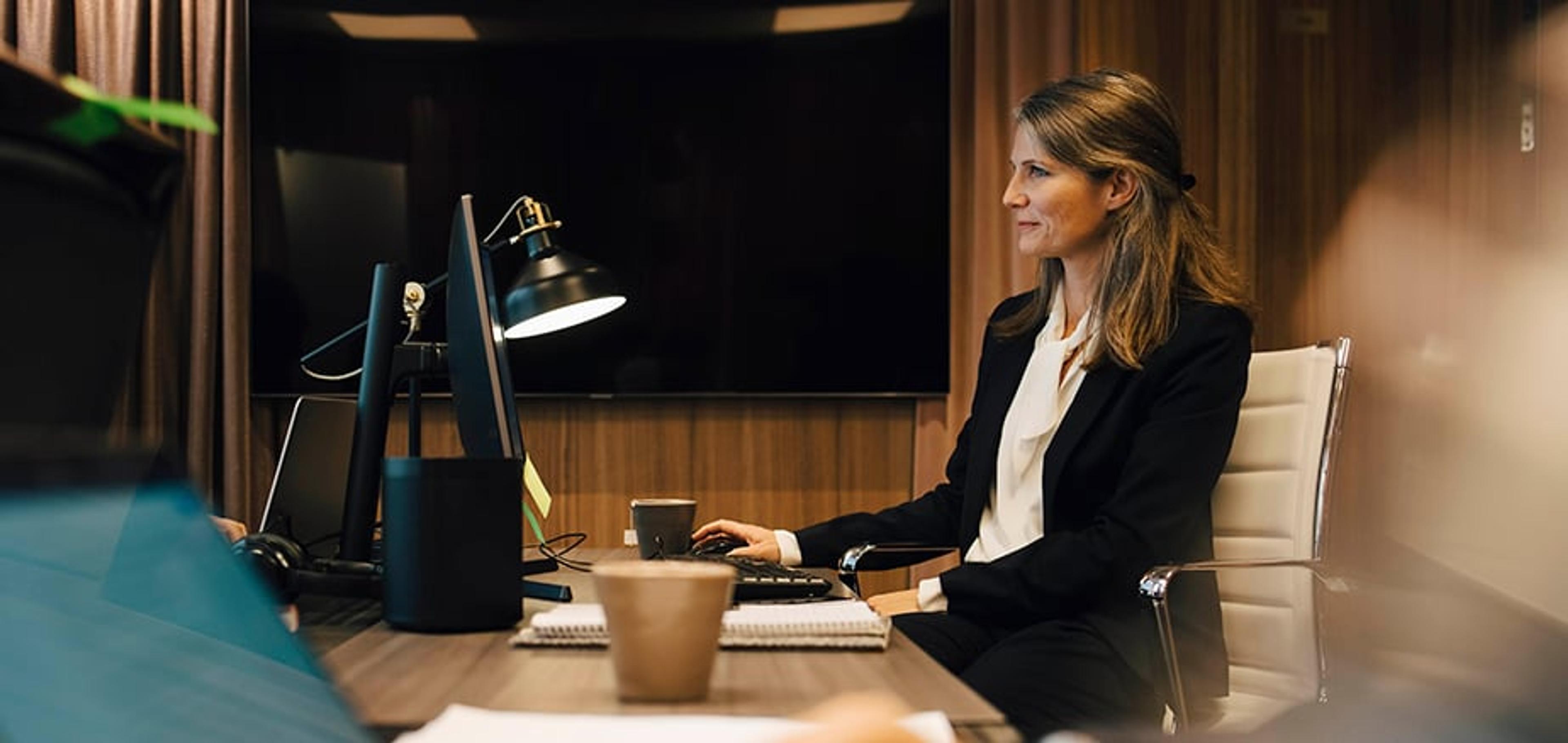 Kvinne i dress sitter ved et skrivebord i et moderne kontor og jobber på en datamaskin, med en stor skjerm og skrivebordslampe foran seg.