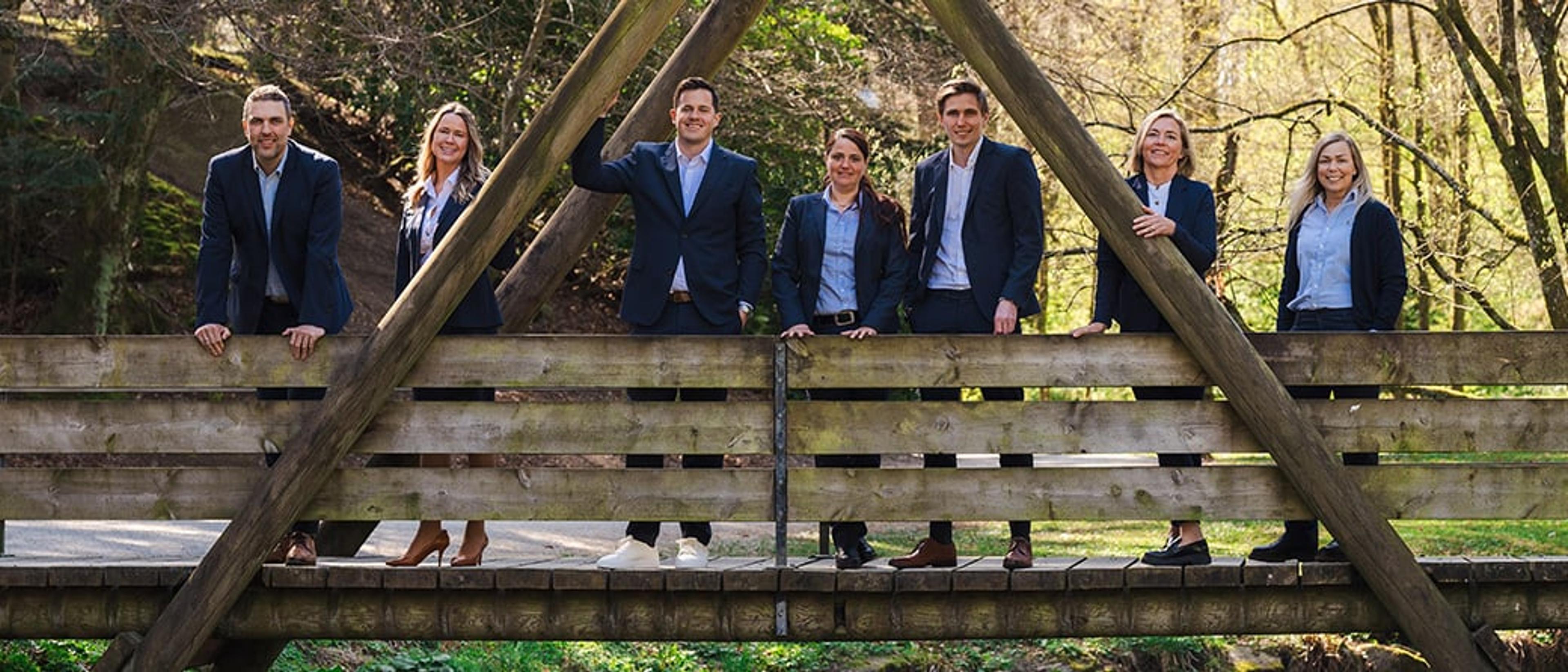 En gruppe på sju personer i formelle klær står på en trebro utendørs, omgitt av grønne trær og vårlys. De smiler mot kameraet i en uformell, profesjonell gruppesituasjon.