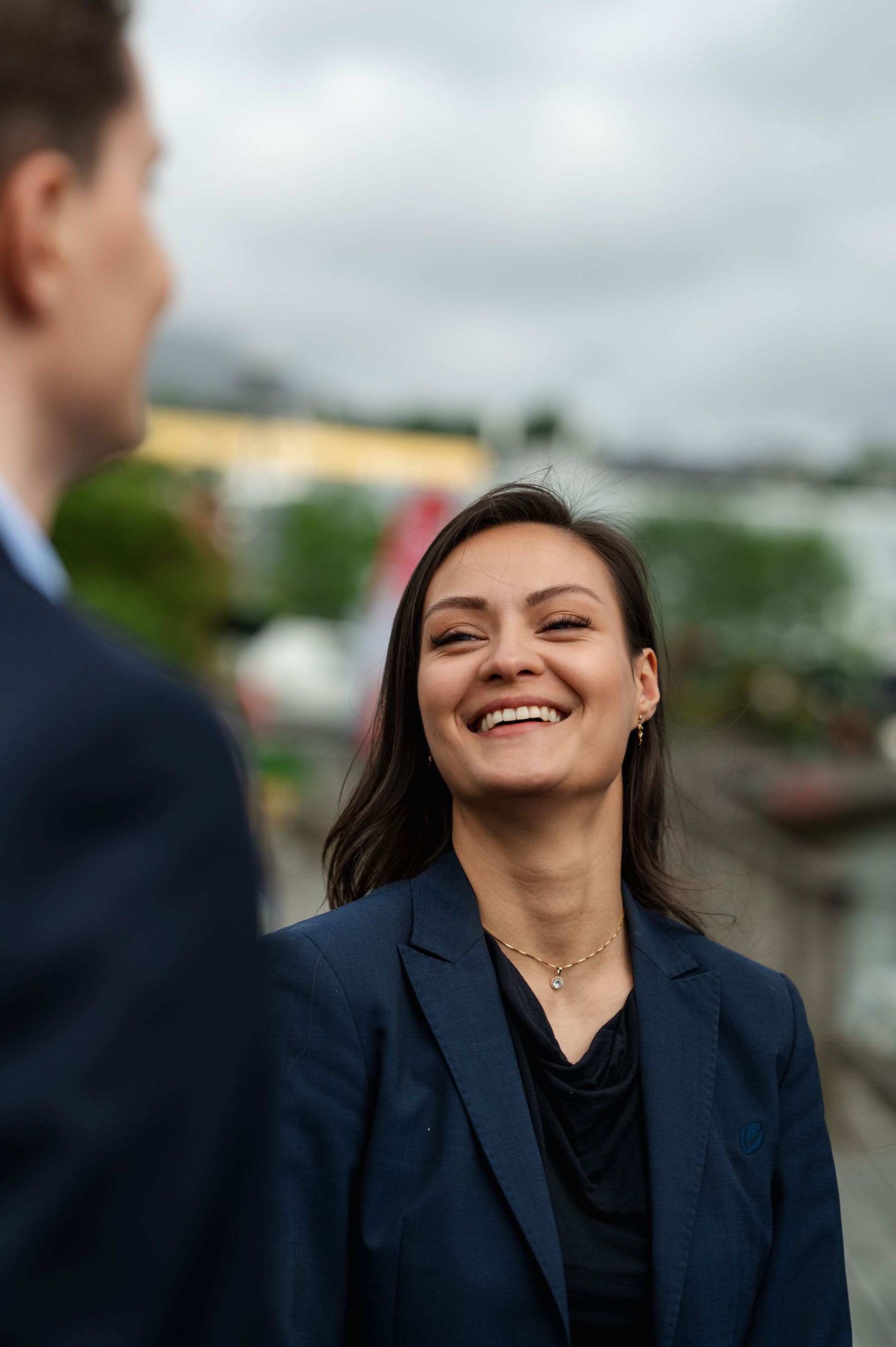 En smilende kvinne i mørk blazer ser opp på en mann mens de snakker sammen utendørs.