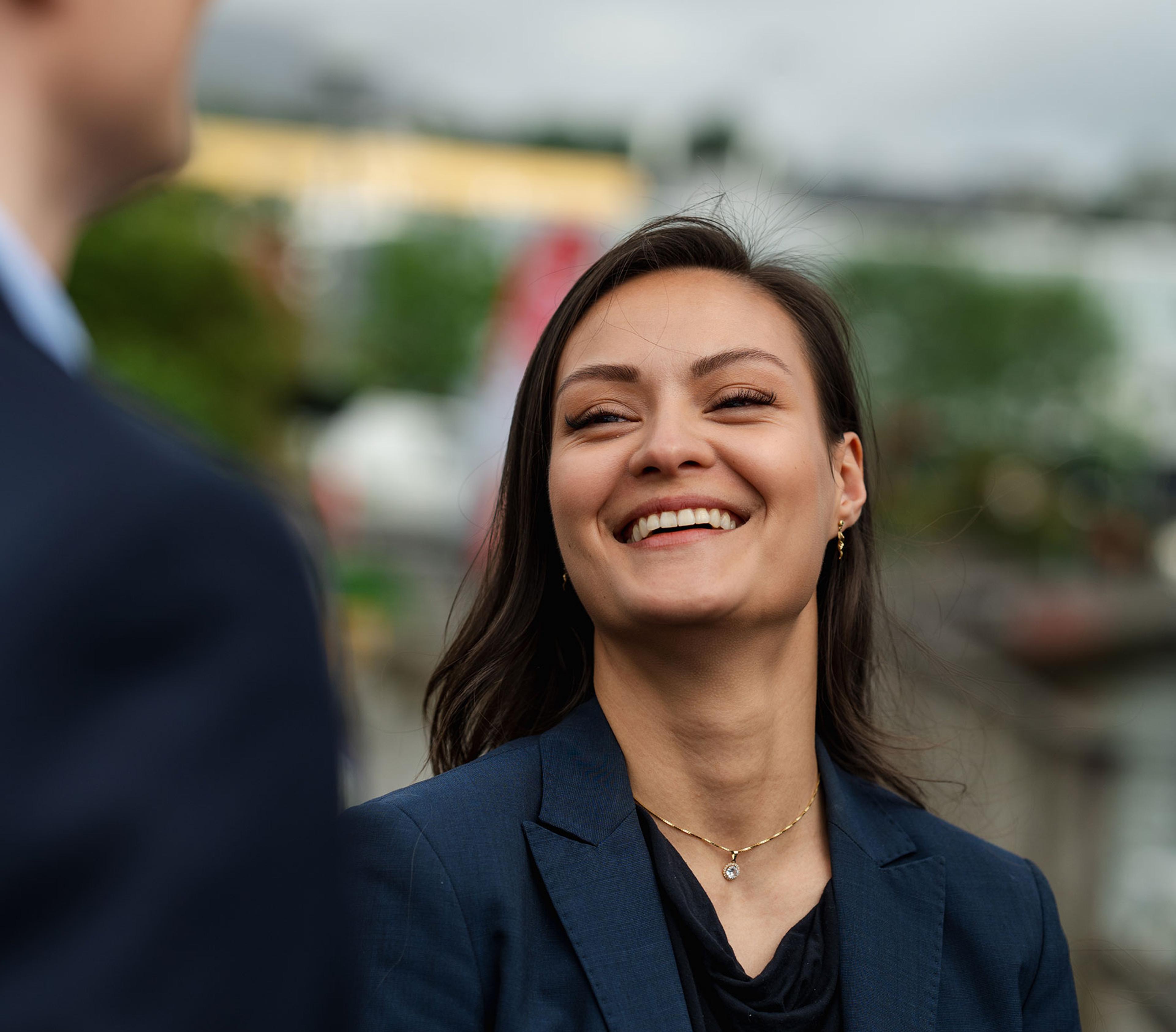 En smilende kvinne i mørk blazer ser opp på en mann mens de snakker sammen utendørs.