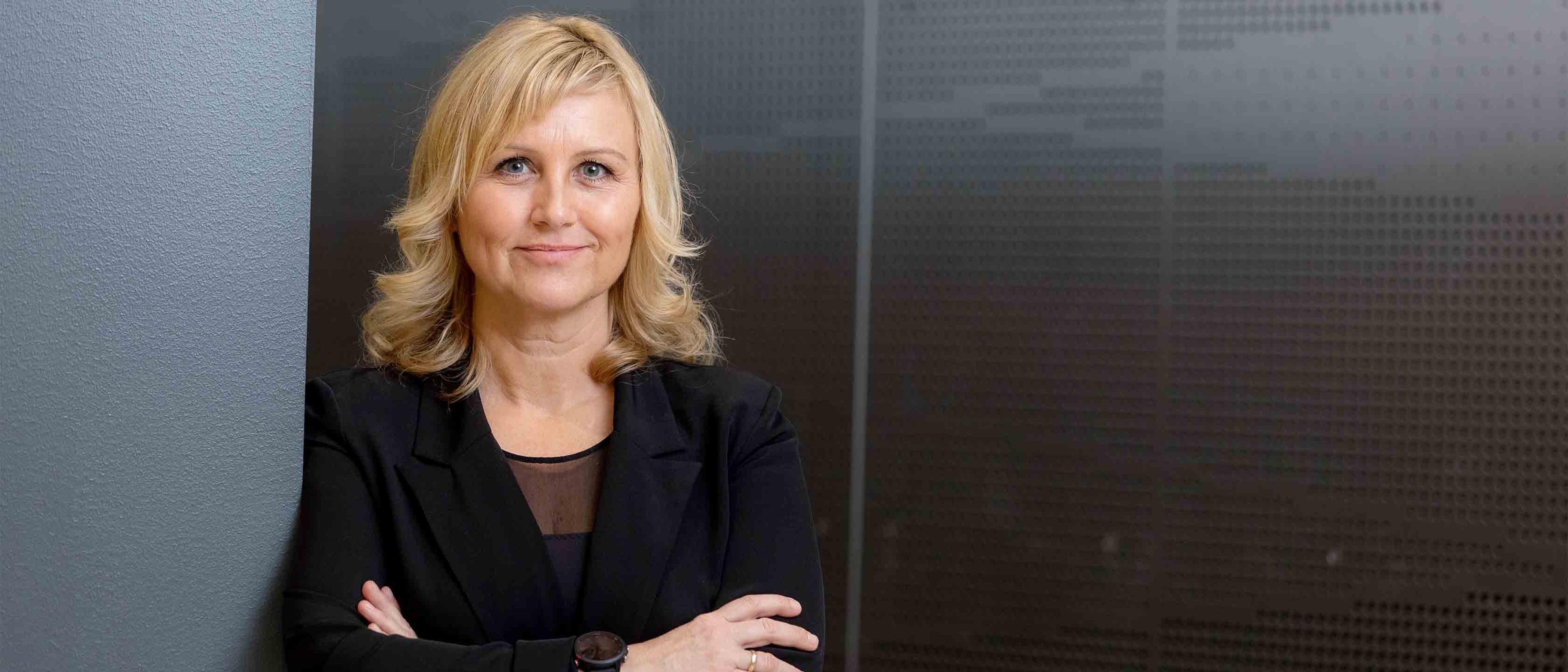 Siren Sundland i svart blazer står med armene i kors og smiler svakt foran en mørk, mønstret vegg.