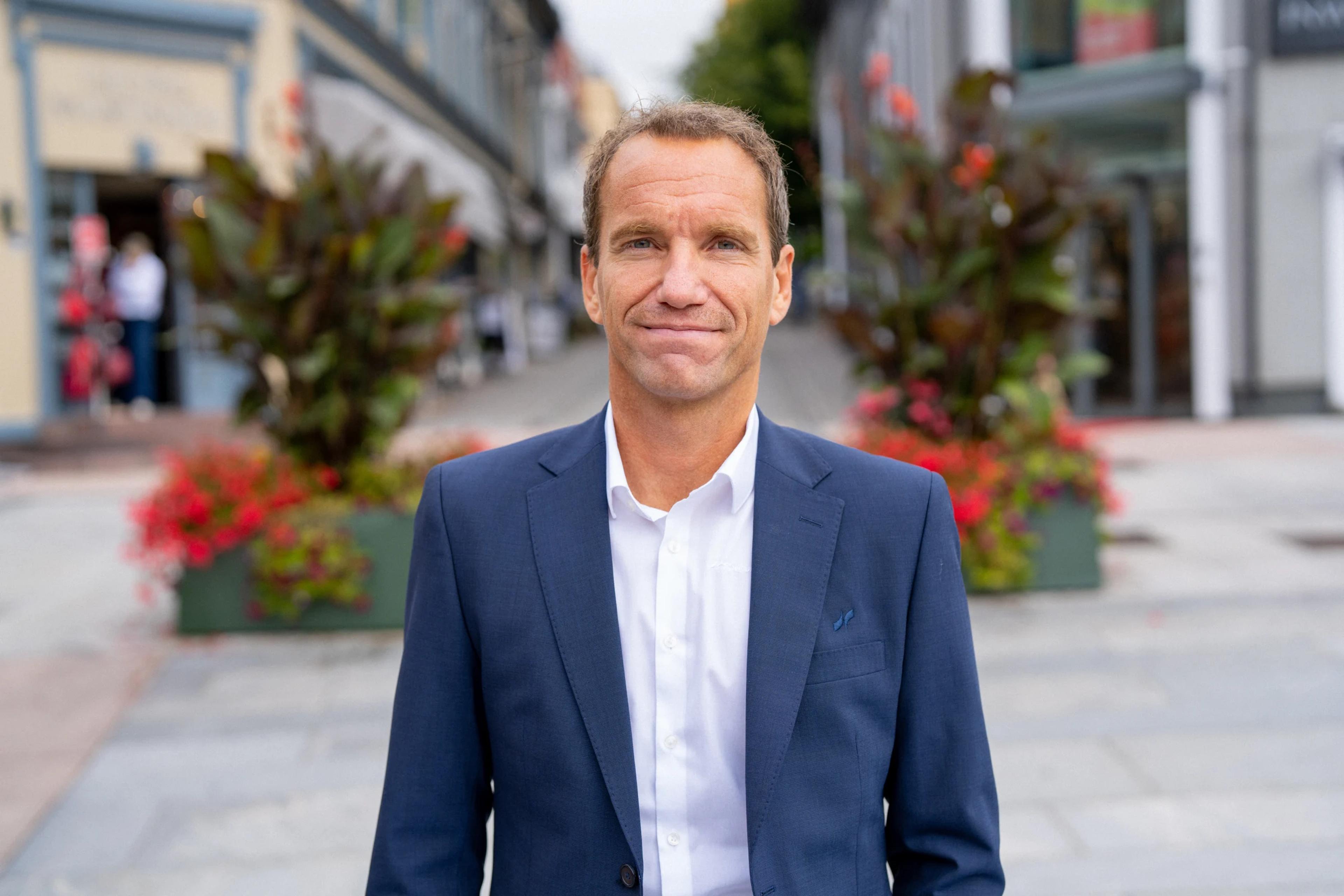 En mann i mørkeblå dressjakke og hvit skjorte står i en gågate med blomsterkasser og butikker i bakgrunnen.
