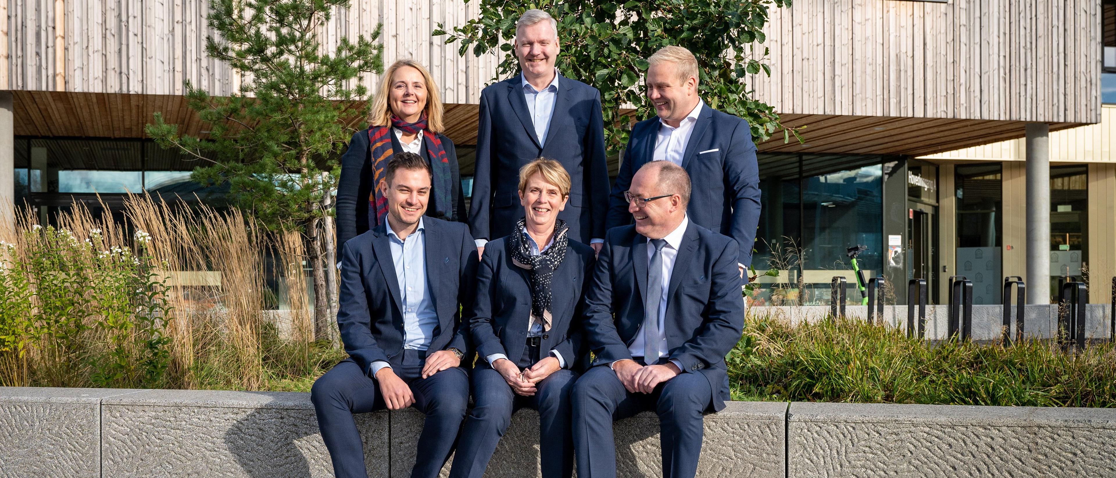 En gruppe på seks forretningsfolk i dress sitter og står utendørs foran en moderne trebygning, smilende mot kameraet i solskinn.